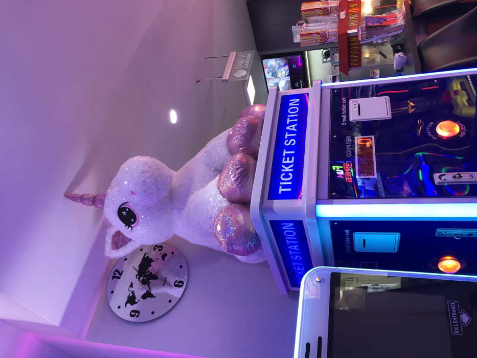 This image, taken in Iraklio, Greece, depicts an indoor entertainment venue, likely an arcade, with a distinct purple and pink lighting scheme. The photograph is oriented vertically, making the subjects appear sideways.
A large, sparkling white unicorn plush toy or statue with iridescent pink hooves and horn is prominently featured, seemingly mounted on a wall or ceiling. Its eye has a black iris with a white star. Below the unicorn, a white circular clock with a black world map design displays the numbers 12, 3, 6, and 9.
To the right, two "TICKET STATION" machines are visible, illuminated with bright blue light. Each machine features a "Small ticket exit," a "TICKET COUNTER" displaying "FUV," and an orange "PUSH FOR RECEIPT" button. The foreground shows part of a white smartphone or similar device.
In the background, stacks of "POPCORN" containers are visible, and a hanging sign from the ceiling partially reads "TAMEAD CAS".