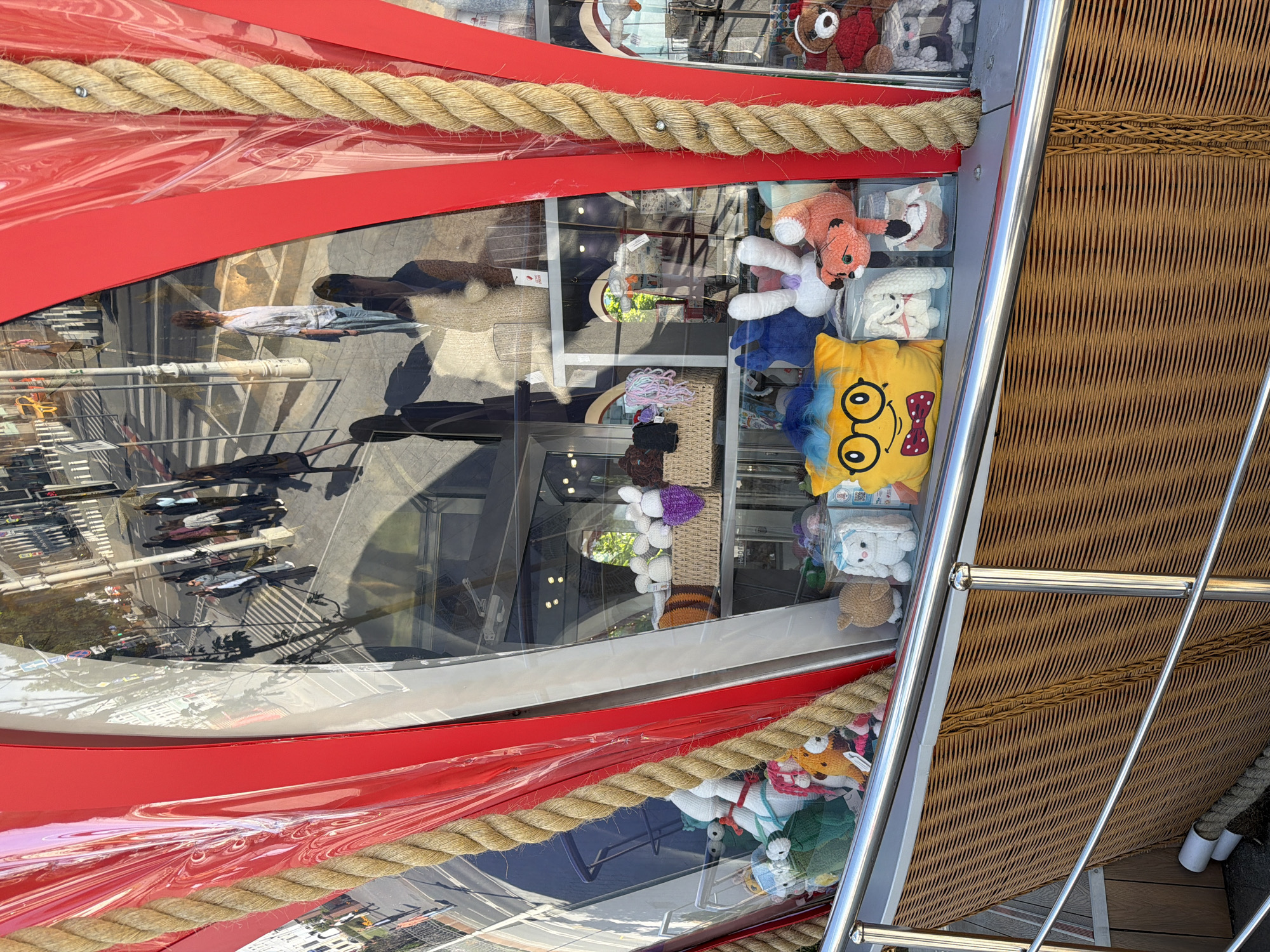 The image shows a close-up view of a shop window display, likely in Moscow, Russia, given the architectural style visible in the reflection. The display features a variety of knitted stuffed animals. In the foreground, a yellow pillow-shaped stuffed toy with blue hair, glasses, and a red bow tie is prominently placed. To its right, a white stuffed rabbit in a clear box sits next to an orange stuffed fox. Further to the right, a brown teddy bear and other stuffed animals are visible on shelves. The window display is framed by red panels and a thick, braided rope. Reflections in the glass show a street scene with pedestrians, buildings, and traffic. The shop window is set into a structure with a wicker-like texture at the bottom.