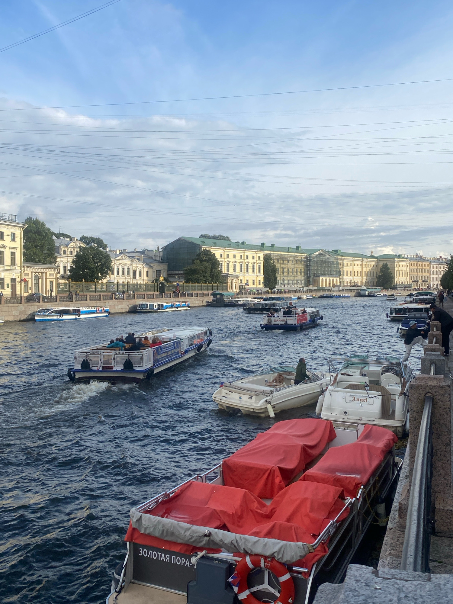 This is an eye-level, wide shot of the Griboyedov Canal in Saint Petersburg, Russia. The canal is filled with several tour boats, some of which are covered with red tarps. A few people are visible on the boats and on the embankment. The buildings lining the canal are typical of Saint Petersburg's architectural style, with ornate facades and classical detailing. The sky is cloudy, and the water is choppy, suggesting a breezy day. The scene conveys a sense of a busy tourist area, with the boats likely offering scenic tours of the city.