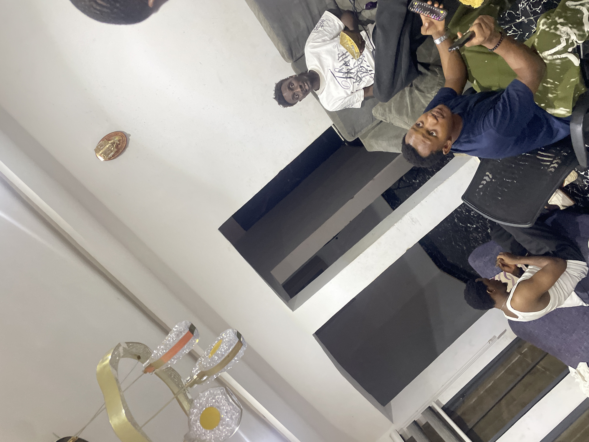 The interior scene appears to be a living room, with three men present. One man is sitting on a grey sofa, wearing a white t-shirt. Another man is sitting in an office chair, wearing a dark blue t-shirt and holding a remote control and cell phone in his hands. A third man is reclined on a purple couch, wearing a white tank top. There are windows visible in the room. A modern chandelier hangs from the ceiling. A religious object is on the wall. The location is likely Abuja, Nigeria.