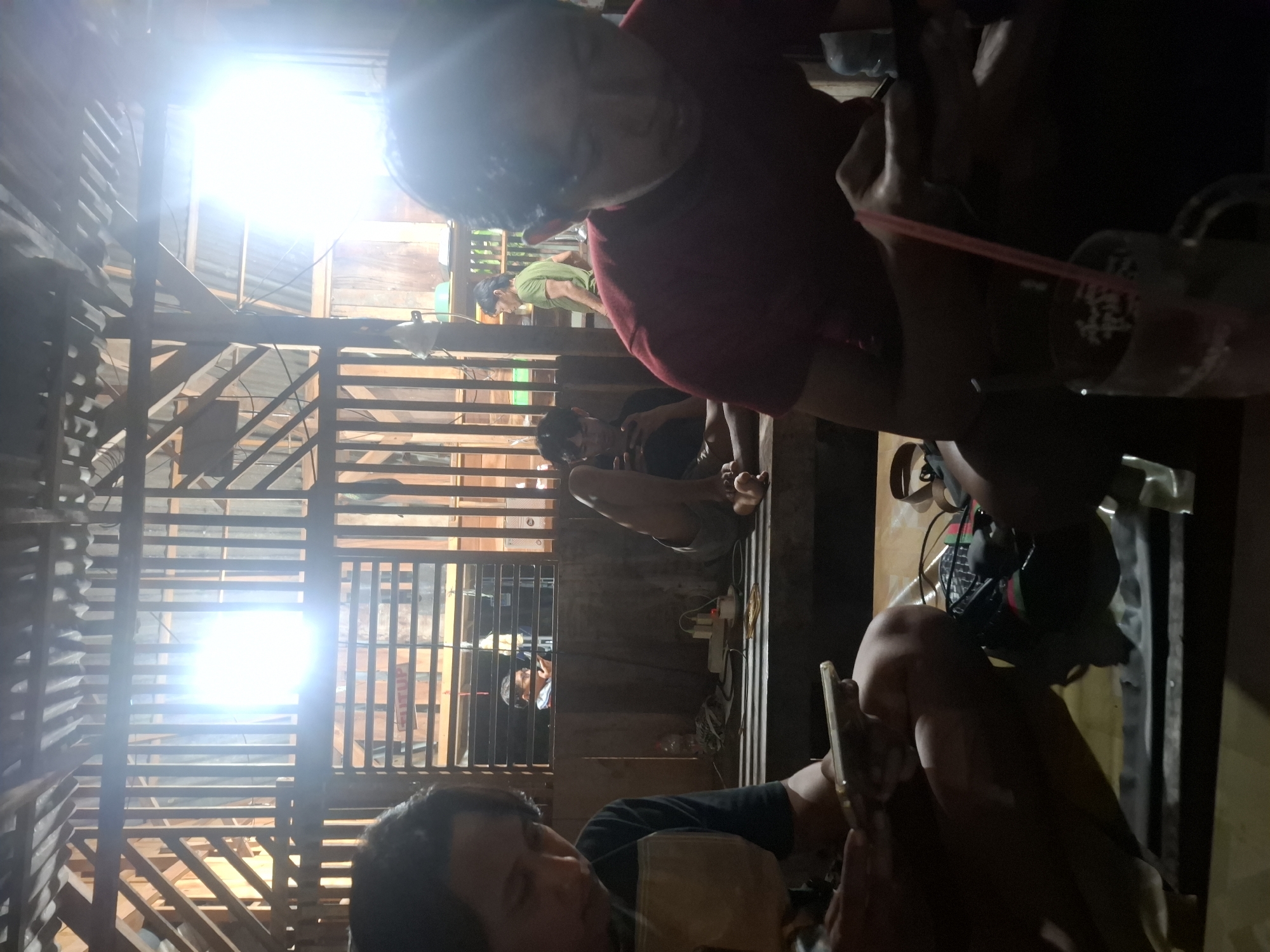 The image depicts a dimly lit interior scene, likely a rustic wooden structure in Bulubrangsi, Indonesia. In the foreground, two individuals are seated, engrossed in their mobile phones. The person on the left, wearing a dark shirt with a light-colored collar, holds a phone in their hands. To the right, another individual in a red shirt is also looking down at a phone. Their posture suggests a casual gathering. In the middle ground, a third person is reclining with their legs up, also appearing to use a phone. Further back, several other figures are visible, including one in a green shirt standing near a window with horizontal wooden slats. Behind the figures, the structure features wooden walls and a corrugated roof, with bright lights creating a strong glare in the upper portion of the image. Several objects are scattered on the wooden surfaces, including a bottle, a power strip with cords, and a bag. The overall impression is one of an informal social gathering at night, possibly in a dwelling or community space.