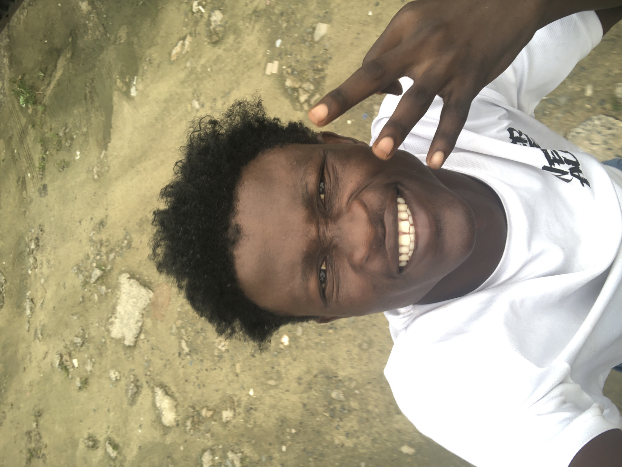 The image shows a close-up, high-angle selfie of a young Black male taken outdoors. He is smiling broadly with his teeth showing and making a peace sign with his right hand, with his index and middle fingers extended. He is wearing a white t-shirt. The background is a dirt or gravel surface with scattered debris. The specific location in Ado, Nigeria, cannot be determined from the visual information provided.
