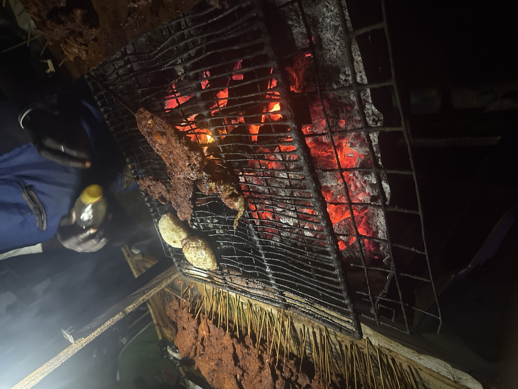 The image depicts a nighttime scene of an outdoor grilling operation, likely in Galadimawa, Nigeria. The central activity involves the preparation of food over a vibrant charcoal fire.
On the left side of the frame, a person's dark-skinned hand, clad in a blue sleeve, is visible holding a small, yellow-capped bottle, possibly containing oil or a spice blend for basting. The hand is positioned near a robust metal grill grate, under which bright red, glowing charcoal embers provide intense heat and the primary light source for the scene.
Cooking on the grill are several skewers of seasoned meat, typical of "suya," a popular West African grilled dish. Their surfaces appear well-cooked and rich brown. Additionally, two lighter-colored, rounded food items, possibly potatoes or similar accompaniments, are roasting alongside the meat. Below the main grill, a wooden surface holds a substantial quantity of raw, reddish-brown marinated meat skewers, arranged on sticks, indicating ongoing preparation or a busy stall.
Smoke subtly rises from the cooking area, illuminated by the hot coals. The rustic setup suggests a street food vendor or an informal outdoor eatery, operating in the dark, with the fire's glow creating a warm and inviting atmosphere. No visible text is present in the image.