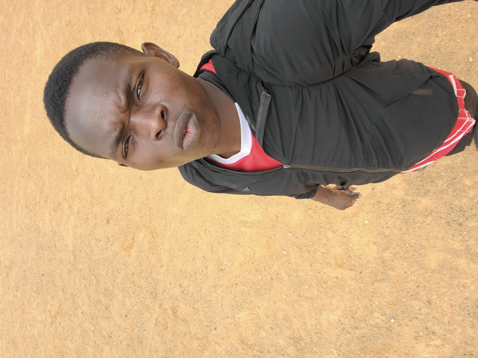 A person with dark skin and short, dark hair is standing on a sandy, tan surface. The person is wearing a red shirt with white trim under a black jacket. The location is Bukuru, Nigeria.
