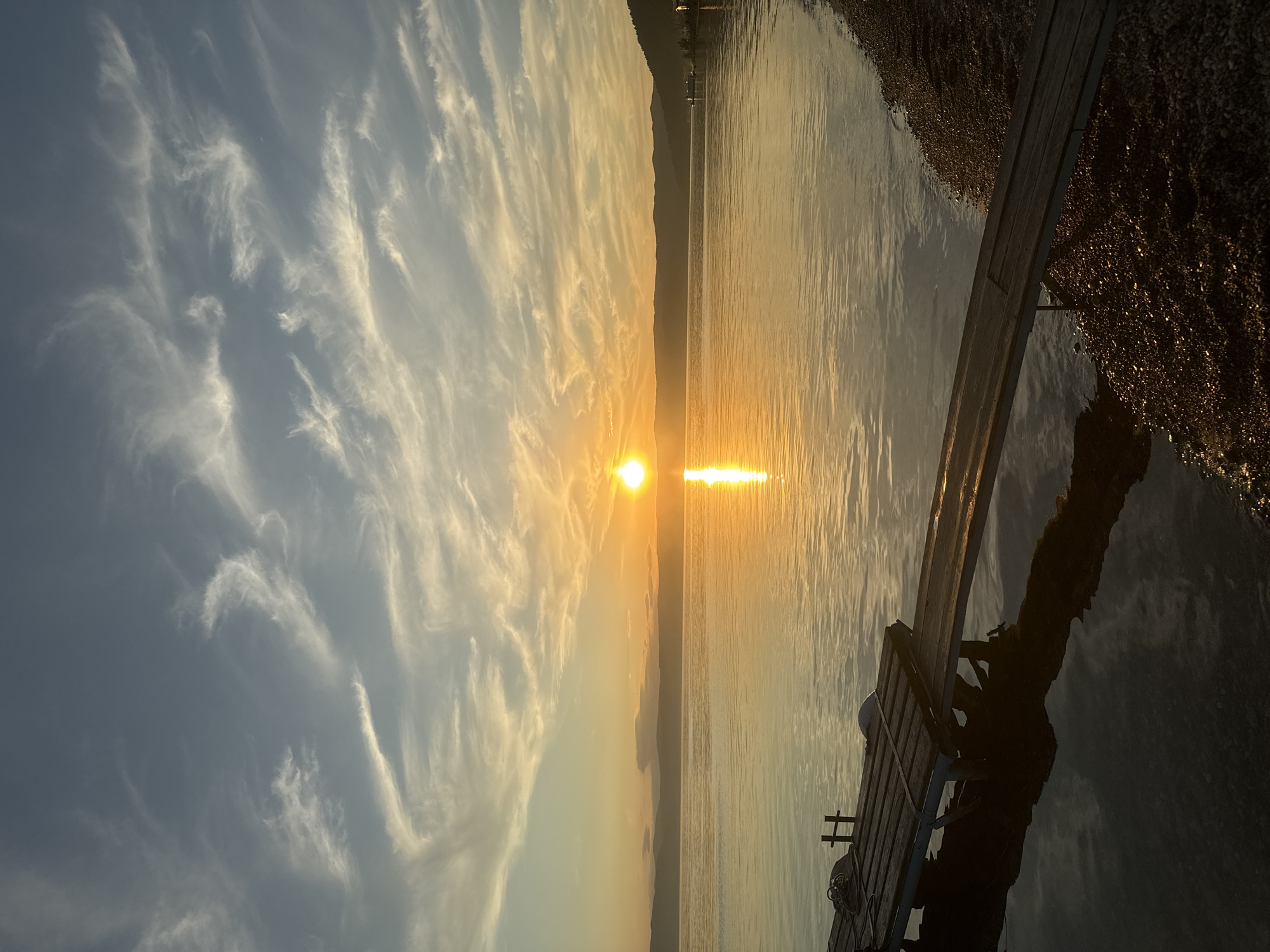 The image captures a serene sunset over a body of water, likely Lake Baikal, given the location of Listvyanka, Russia. A wooden dock extends from the rocky shore into the calm, reflective water. The sun, a bright orb, is setting on the horizon, casting a warm, golden glow that illuminates the sky and its wispy clouds. The water mirrors the sunset, creating a shimmering path of light. Distant, rolling hills form the backdrop. The scene evokes a sense of peace and natural beauty.