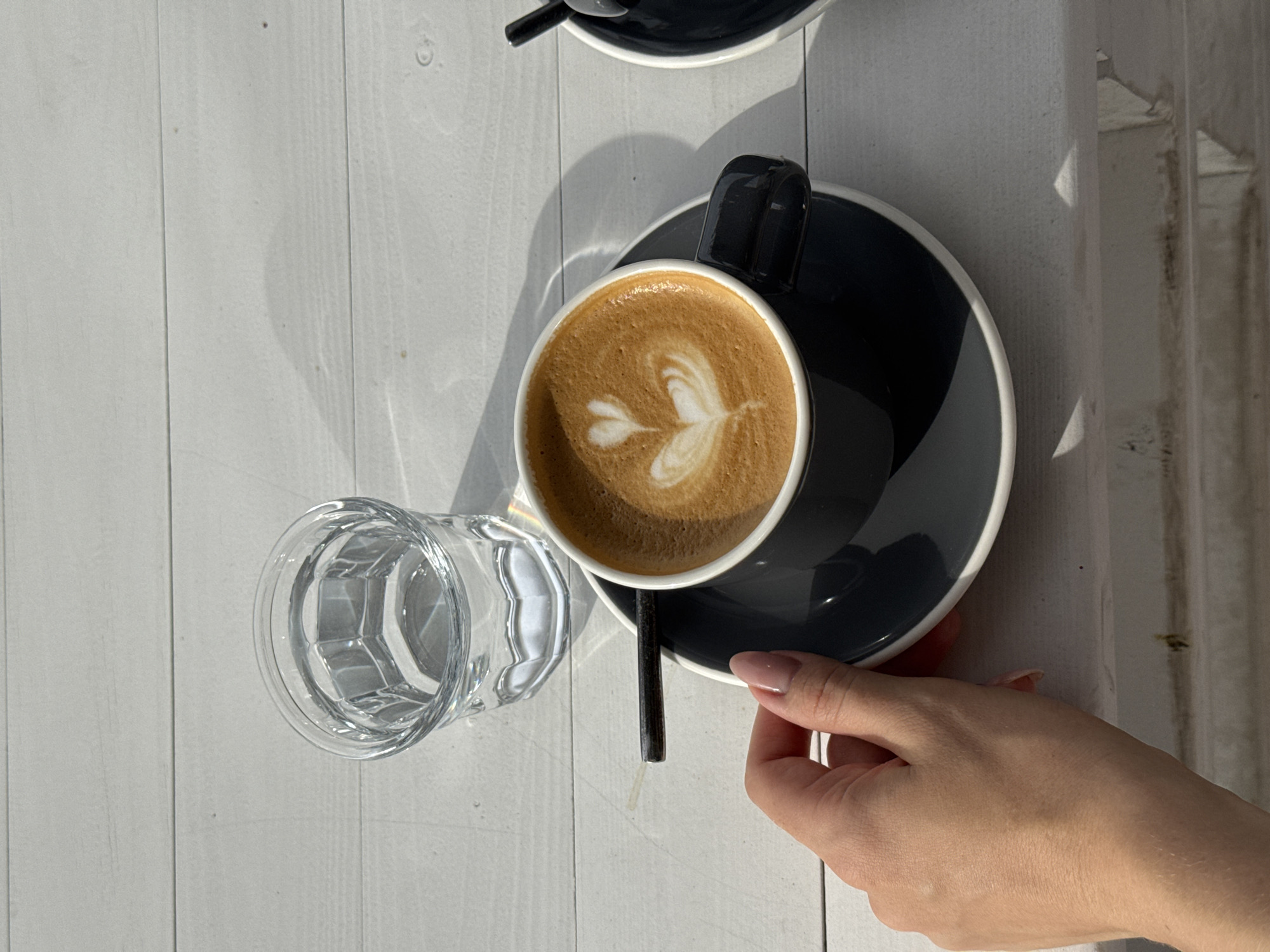 The image captures a scene on a white wooden table in Unknown, Unknown. A cup of coffee, adorned with latte art, rests on a black saucer. Next to the coffee is a clear glass filled with water. A hand is visible, partially holding the edge of the saucer. Another cup, partially visible, is placed at the top of the image. Sunlight casts shadows across the table, creating a contrast between light and shade.