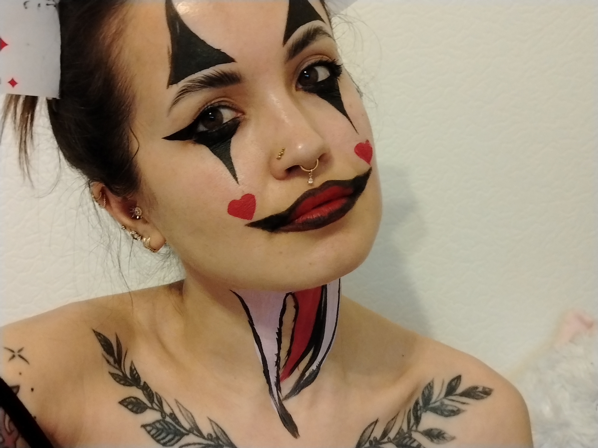 A young woman poses for a close-up photograph. She is facing the camera at an angle with her head slightly tilted. She is wearing clown-like face paint, with black triangular shapes above and below her eyes, red hearts on her cheeks, and black paint extending from the corners of her mouth. She has dark hair pulled back in a bun with a playing card (possibly a heart) attached. She has multiple piercings in her left ear and two piercings in her nose. She has tattoos on her chest and shoulder, one appearing to be a floral design. Her neck has painted designs in black, red, and white. The background is a textured white wall.
I cannot tell you the location of this image.