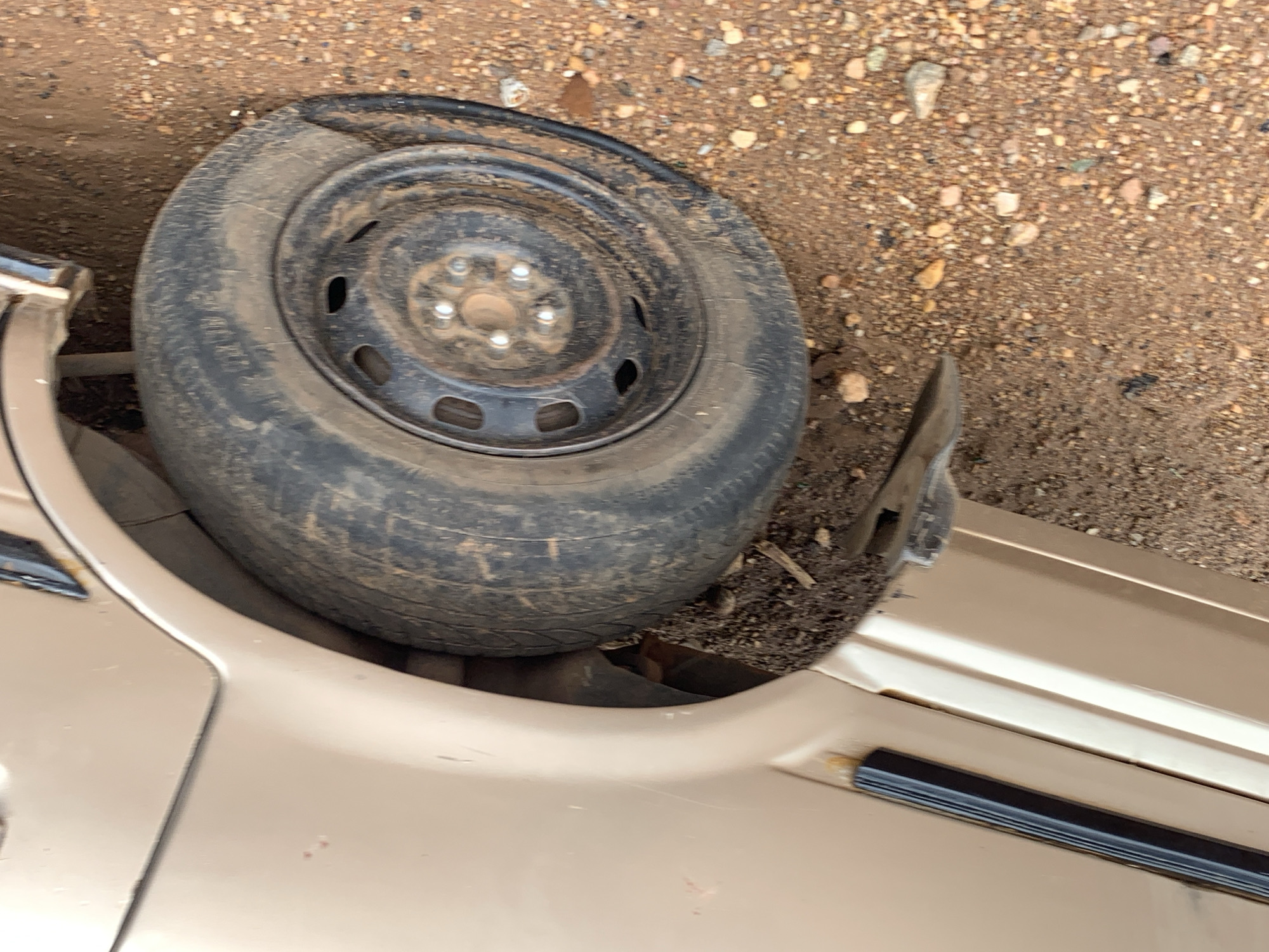 The image shows a damaged gold-colored vehicle with a flat tire lying on the ground near the car. The tire is black and dirty, with a steel rim. The surrounding ground is a mix of dirt and small rocks. There is no evidence indicating the location to be Rafin Par, Nigeria. No people are visible.