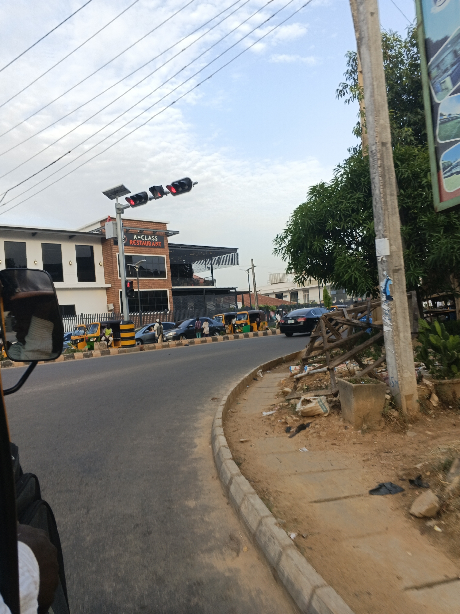 The image depicts a street scene in Kabala, Nigeria, from the perspective of someone riding in a vehicle (possibly a tricycle, judging by the partially visible mirror and frame). The road curves to the left. On the left side of the road is a two-story building with a sign that reads "A-CLASS RESTAURANT." A solar-powered traffic light is visible above the restaurant. Several vehicles, including cars and yellow and green tricycles (Keke Napeps), are parked or moving along the street. On the right side of the road is a curb, followed by a dirt and concrete sidewalk with trash and debris. A utility pole is present on the right edge of the frame, partially obscured by foliage and what appears to be makeshift wooden support. The sky is partly cloudy.