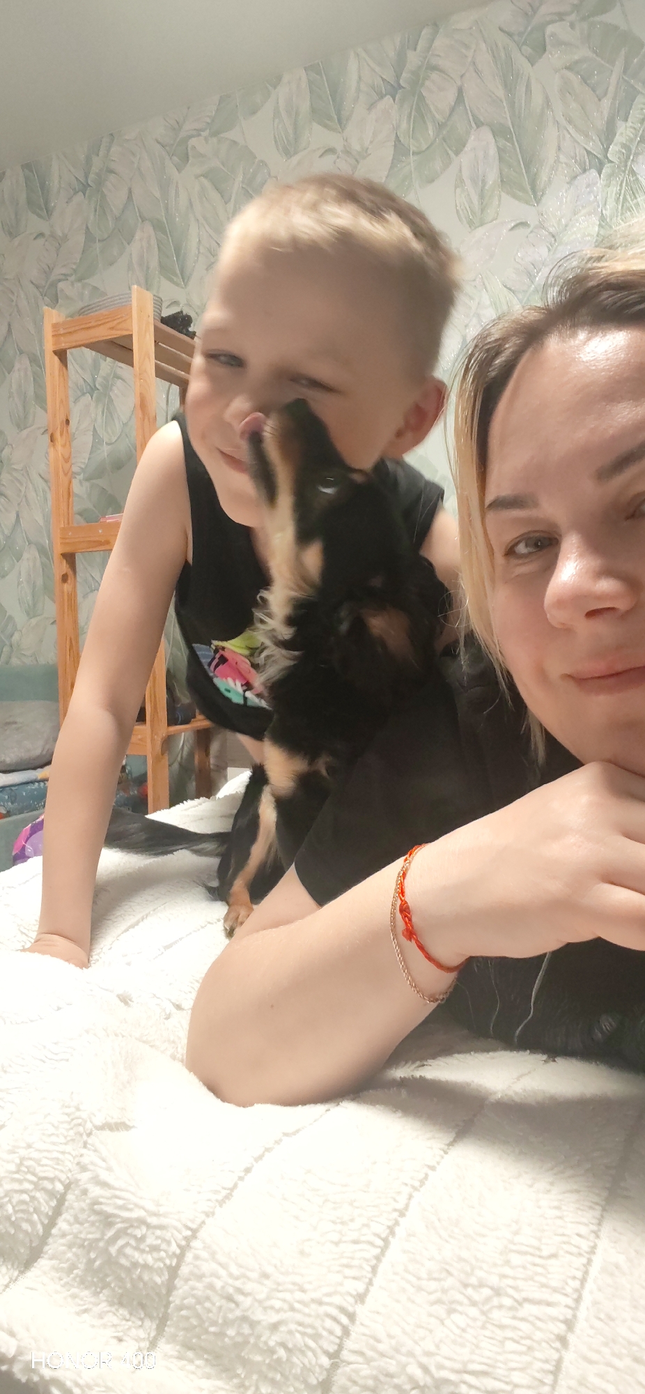 A woman, a child, and a dog are in a room with patterned wallpaper and a wooden shelf. The child is being licked by the dog. The woman is looking at the camera, with her arm resting on a white textured surface. There is an inscription "HONOR 100" at the bottom. The location in the city of Noviy, Russia, is not discernible from the image.