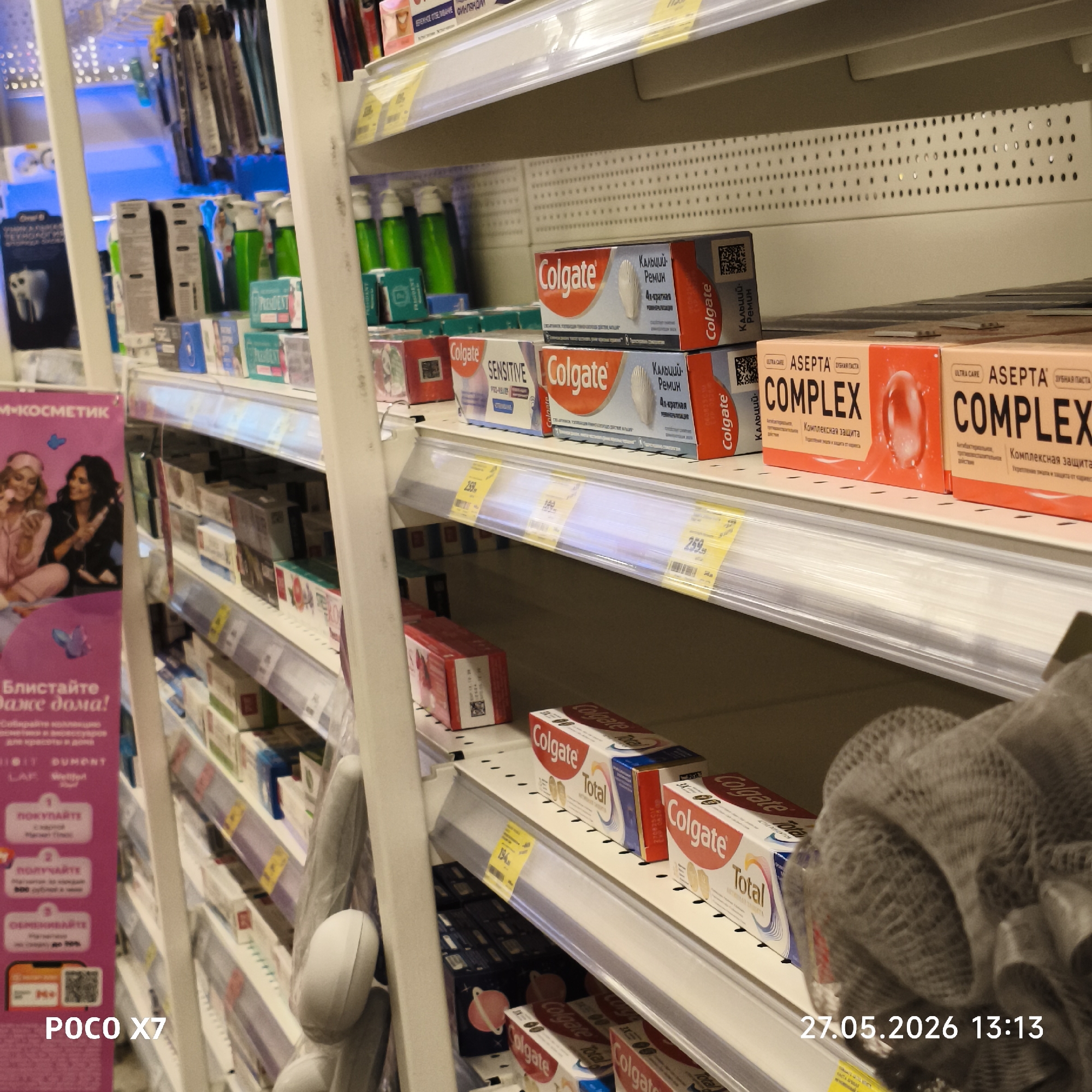 This image shows shelves in a store, likely in Chaikovsky, Russia, filled with various oral hygiene products. Brands such as Colgate and Asepta are visible. The products are displayed on multiple shelves with price tags underneath. A display advertisement is visible on the left side of the frame. The photograph was taken on May 27, 2026 at 13:13 local time with a POCO X7 smartphone.