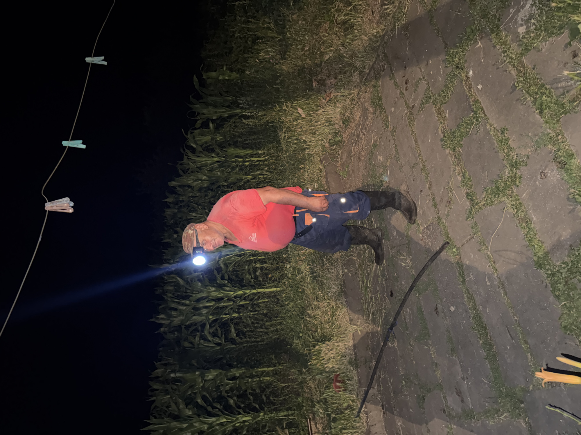 A man stands in a paved area at night, illuminated by a headlamp. He is wearing a red shirt, blue pants with orange accents, and black boots. A cornfield is visible behind him, and a clothesline with clothespins is strung across the top of the image. The location is in Tandzia, Georgia.