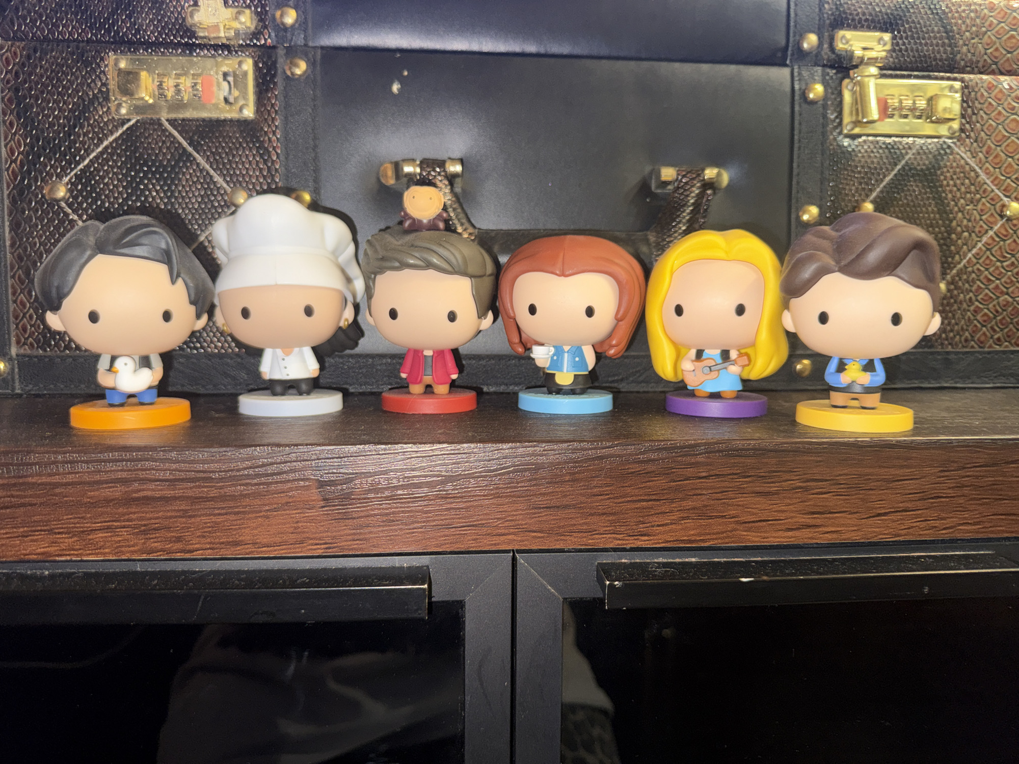 A group of six stylized figurine toys, each depicting a character from the television show *Friends*, are arranged in a line on a wooden shelf. The figurines have oversized heads and simple features, characteristic of collectible vinyl figures. The background is dominated by a large, dark-colored trunk with brass accents and combination locks. The shelf is positioned above two black cabinet doors. The figurines are, from left to right: Joey Tribbiani wearing a white shirt and holding a rubber duck; Monica Geller in a chef's outfit; Chandler Bing in a red jacket; Phoebe Buffay holding a coffee cup; Rachel Green playing a ukulele; and Ross Geller holding a rubber duck. There is no indication of the location in Odesa, Ukraine.