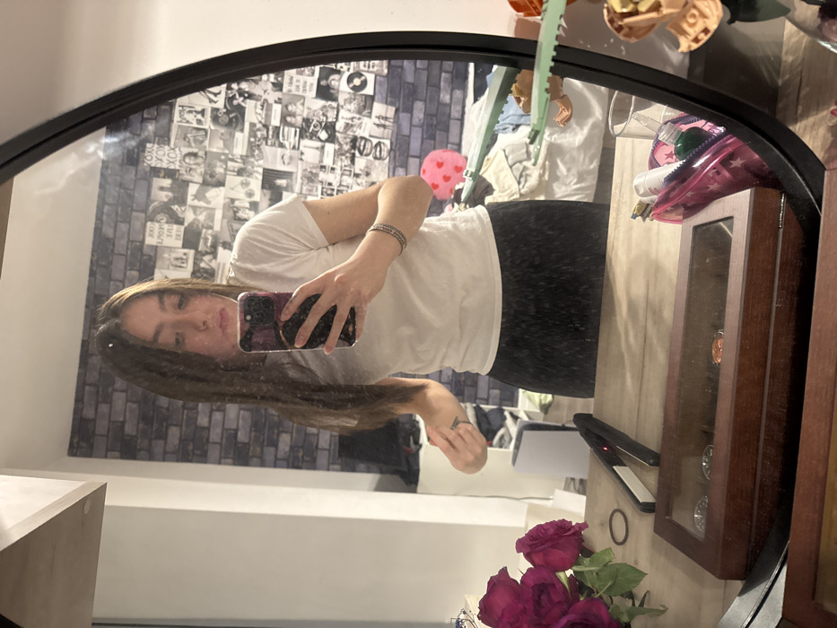A person stands in front of a round mirror, holding a phone to take a selfie. They are wearing a white t-shirt and dark pants. The background includes a wall decorated with many small pictures, a bed, and various items on a surface including red roses, a hair straightener, and a wooden display case. The reflection in the mirror reveals a casual indoor setting.