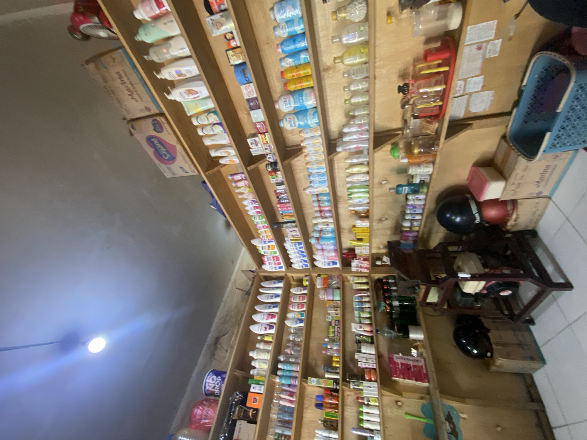 The image shows the interior of a small convenience store or minimarket in Ta', Indonesia. The store is filled with shelves packed with various consumer products, primarily toiletries, beauty products, and cleaning supplies. The shelves are made of light-colored wood and are organized from floor to ceiling.
On the shelves, one can see a wide array of bottled products, including lotions, shampoos, soaps, and sanitizers. Many of the bottles are white, pink, blue, and green. There are also boxes of products, some with branding visible, such as "Marina" and "Sayang". Small jars and containers are also present, holding various items.
In the foreground and on the floor, there are several objects: two black helmets, one on top of a cardboard box, and another on a different surface. A red chair-like structure with a mechanical component is positioned in the center-right of the image. Next to it is a blue plastic basket. There are also some cardboard boxes on the floor.
The lighting in the store appears to be a mix of overhead fluorescent lights and possibly a single bright bulb visible on the ceiling. The walls are a light color, and the ceiling shows some signs of wear. The overall impression is of a cluttered but functional retail space. The presence of the "Marina" brand suggests the store is likely located in Indonesia, a country where this brand is common. The specific location in "Ta'" is a detail that would require external information to confirm beyond the visual evidence.