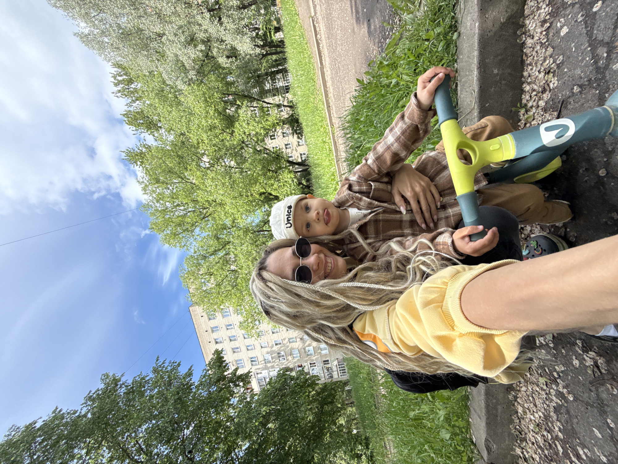 A woman with dreadlocks and sunglasses, wearing a yellow sweater, is taking a selfie with a child wearing a brown plaid shirt and a white cap with the word "Unice" on it. The child is sticking out their tongue. They are sitting on a concrete curb next to some grass. A small green and blue scooter is in front of them. In the background, there are trees, a building with many windows, and a blue sky with some clouds. The location is likely in a residential area of Rusanovo, Russia, based on the style of building.