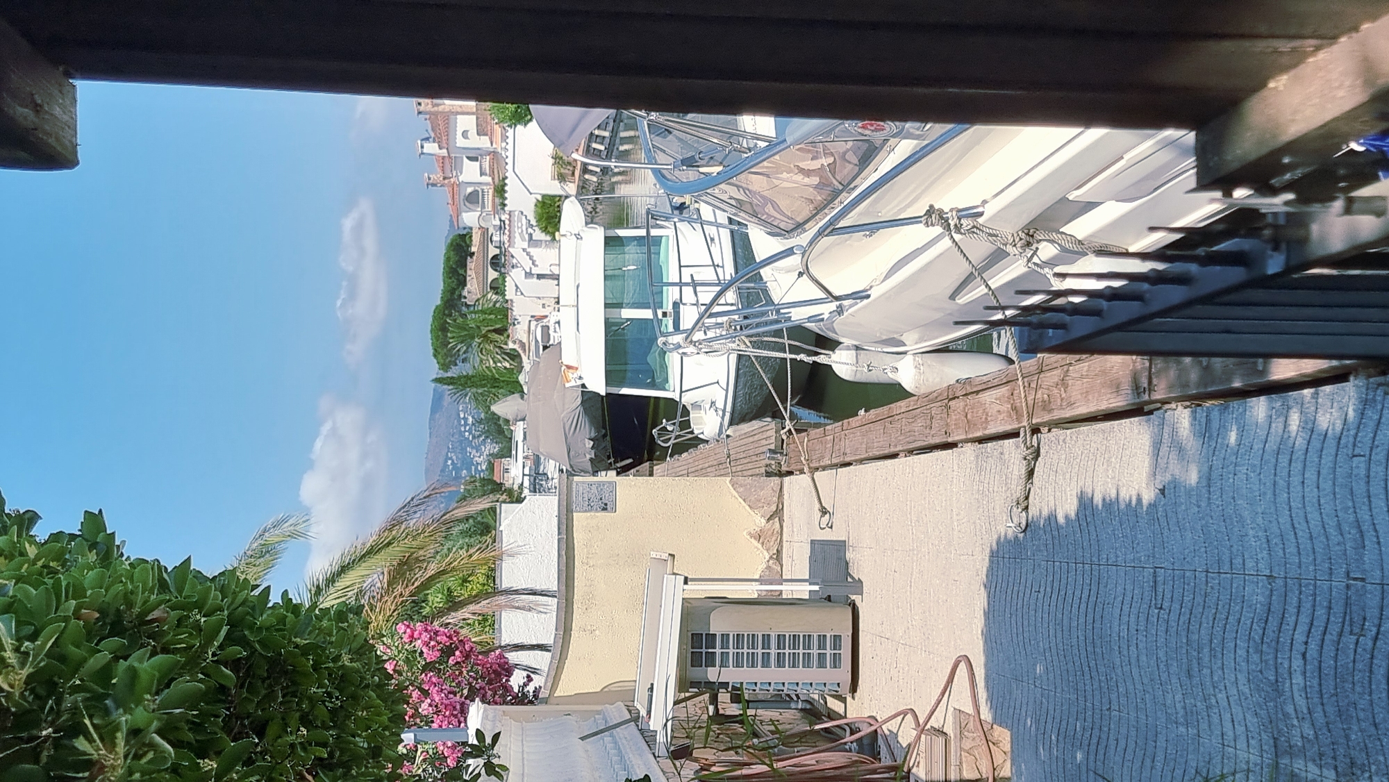 The image captures a view of a canal in Castelló d'Empúries, Spain, likely within the Empuriabrava marina. In the foreground, a paved walkway with a wavy pattern runs along the edge of the canal. To the left, an outdoor air conditioning unit is visible next to a yellow-colored building. Pink flowers and palm fronds are also present on the left side. Several boats are docked in the canal. A white boat with a metal railing is prominently featured on the right, with another boat, partially covered by a grey tarp, docked behind it. The water of the canal is visible between the walkway and the boats. In the background, a row of white buildings with red-tiled roofs, characteristic of coastal Mediterranean architecture, is visible, with a hilly landscape beyond. The sky is clear with a few scattered clouds. The overall scene depicts a sunny day in a waterfront residential or recreational area.