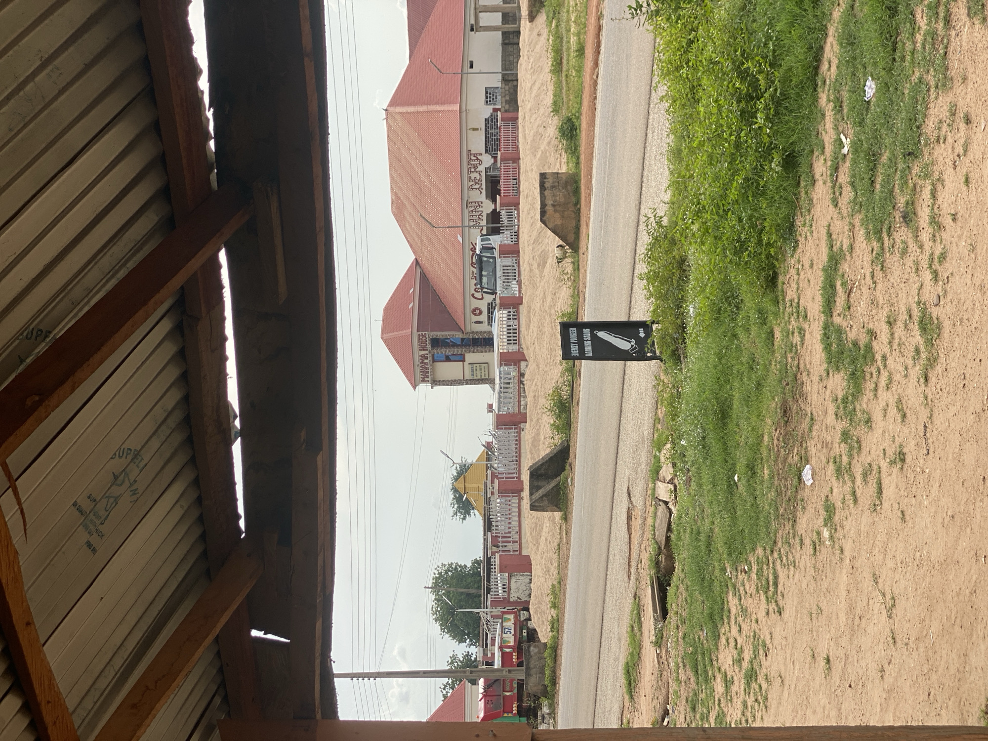 The image depicts a street scene in Bali, Nigeria, with a view through a makeshift roofed structure. In the foreground, the edge of a dirt and grassy area is visible, with some scattered debris including white paper. A road runs horizontally across the mid-ground. On the opposite side of the road, a building complex with red-tiled roofs is prominent. One section of the building has the signage "PANAMA HOUSE" and another section appears to be a "MINI DEPOT." A white truck is parked in front of the depot. A white fence with red posts surrounds a sandy area in front of the buildings. A yellow pyramid-like structure is visible behind the fence. In the foreground, to the left of the road, a black sign with white lettering and an illustration of a barber's clippers stands on the roadside. The sky is overcast.