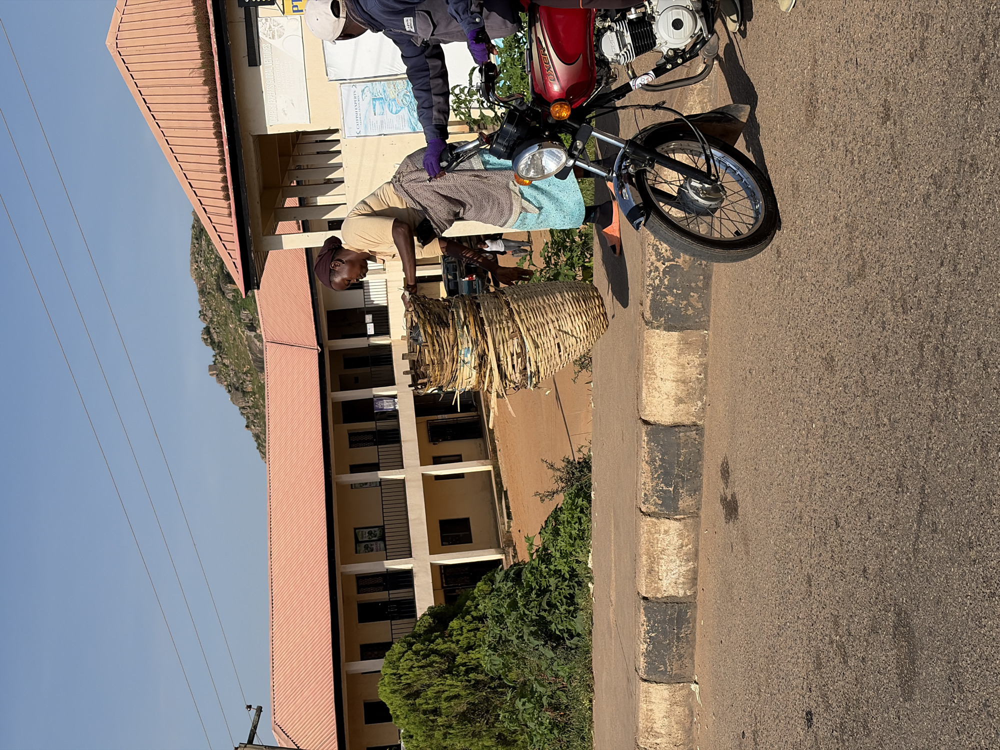 In Gura Top, Nigeria, a woman with dark skin and her hair covered by a dark headscarf, wearing a light brown t-shirt and a blue skirt, stands beside a large stack of woven baskets. She is interacting with another person, whose gloved hands are visible on the handlebars of a red motorcycle. The second person is wearing a dark jacket and a white cap. The scene takes place outdoors, with a building that has a pink roof and cream-colored walls in the background. Power lines are visible against a clear blue sky, and a rocky, mountainous terrain is present in the distance. The foreground shows a paved road and a curb with black and white markings.