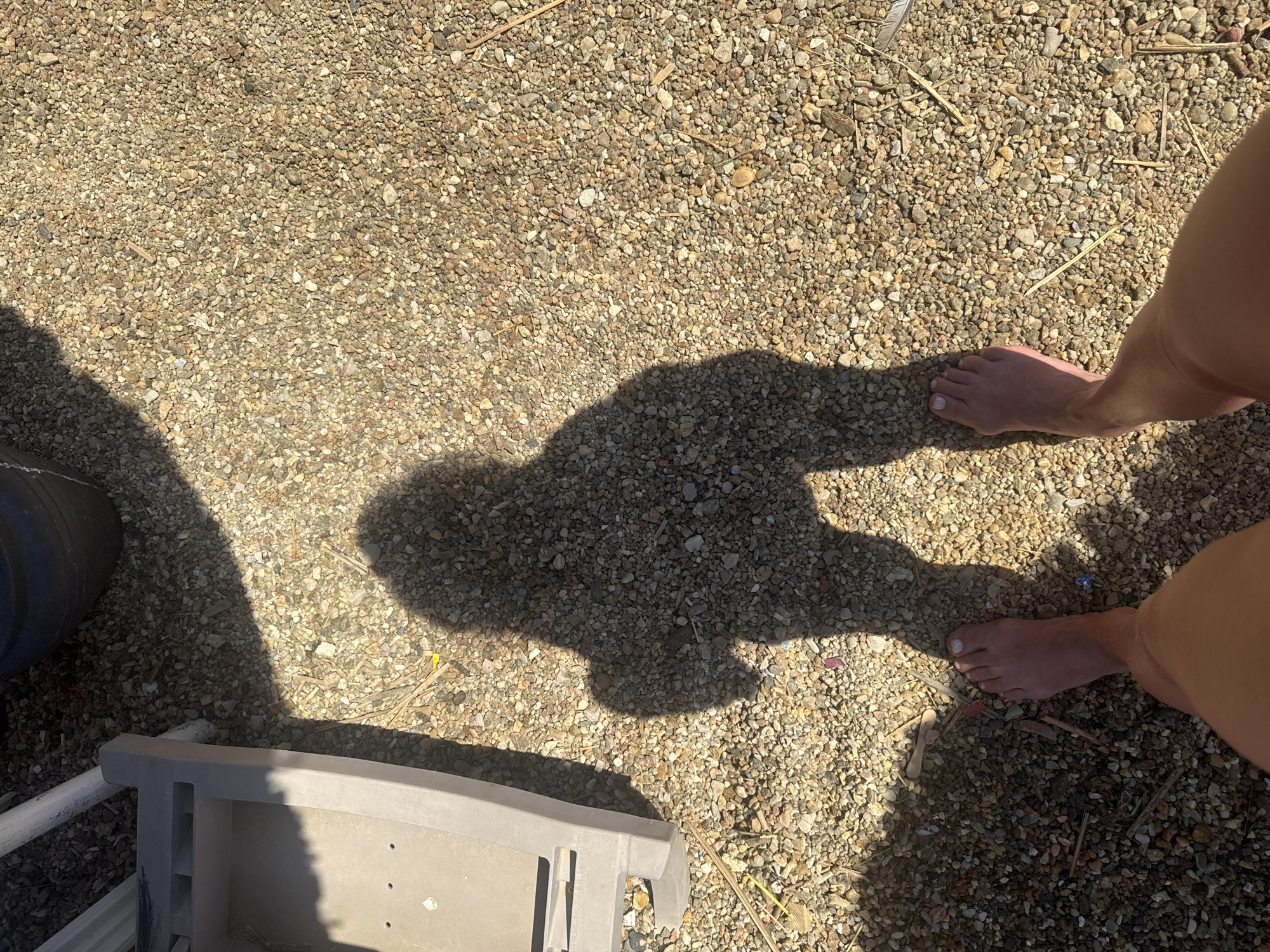 This is a close-up, high-angle shot of a person's bare feet standing on a gravel ground, casting a shadow. The person is wearing shorts. To the left of the frame, a portion of a grey plastic object, possibly a chair or a storage container, is visible. The ground is covered in small, multi-colored pebbles and dry plant debris. The lighting suggests it is a sunny day, with shadows cast by an unseen object or structure. The scene appears to be outdoors, in a dry, possibly arid environment. No specific landmarks or context are provided to pinpoint the exact location within Bodrum, Turkey.