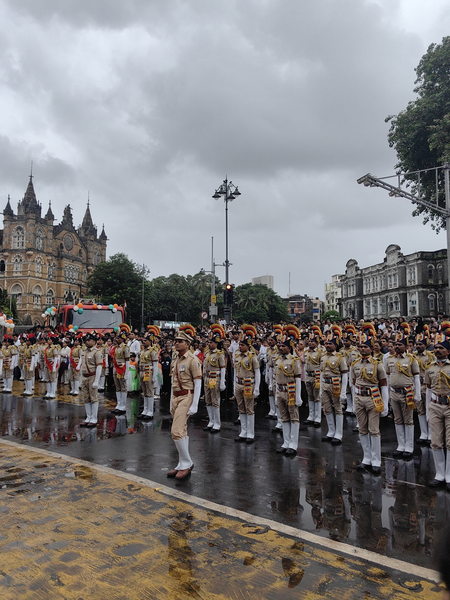 The image captures a scene on a wet street in Mumbai, India, under an overcast sky. A parade is in progress, with a formation of men in ceremonial uniforms standing at attention. The uniforms appear to be light brown trousers, white socks, brown shoes, and tan shirts with ornate collars and epaulets. Many wear distinctive turbans with colorful plumes. In the background, a large, ornate building, possibly a historical landmark, stands prominently. A red fire truck decorated with balloons in the colors of the Indian flag is visible behind the parade participants. A crowd of onlookers lines the street. The wet asphalt reflects the figures and structures, indicating recent rainfall. The overall atmosphere suggests a formal civic event, likely a national celebration.