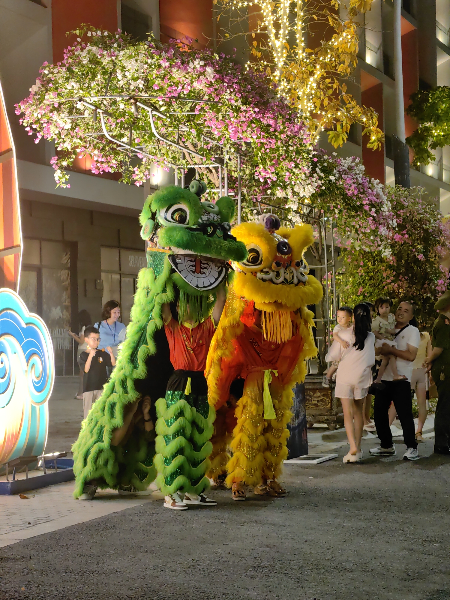 Captured in Bai Chay Ward, Vietnam, the image shows two people performing the traditional Lion Dance. One is wearing a green lion costume, and the other is wearing a yellow one. They are surrounded by spectators, including adults and children. Some of the adults are holding young children. The backdrop features illuminated flora with pink and white blossoms, as well as other decorative light fixtures. The location appears to be a street, and the setting suggests an evening event or celebration.