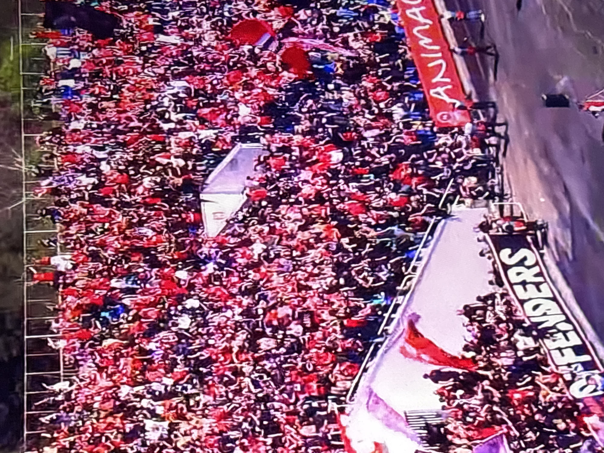 An aerial view of a large crowd, primarily dressed in red, at a sporting event. The crowd is dense and occupies stadium seating next to a paved road. There are some red flags in the crowd. A white banner with "DEFENDERS" printed in black capital letters is visible. A red banner with the word "ANIMA" (partially obscured) is visible further along the road. The location is most likely in the city of Sofia, Bulgaria.