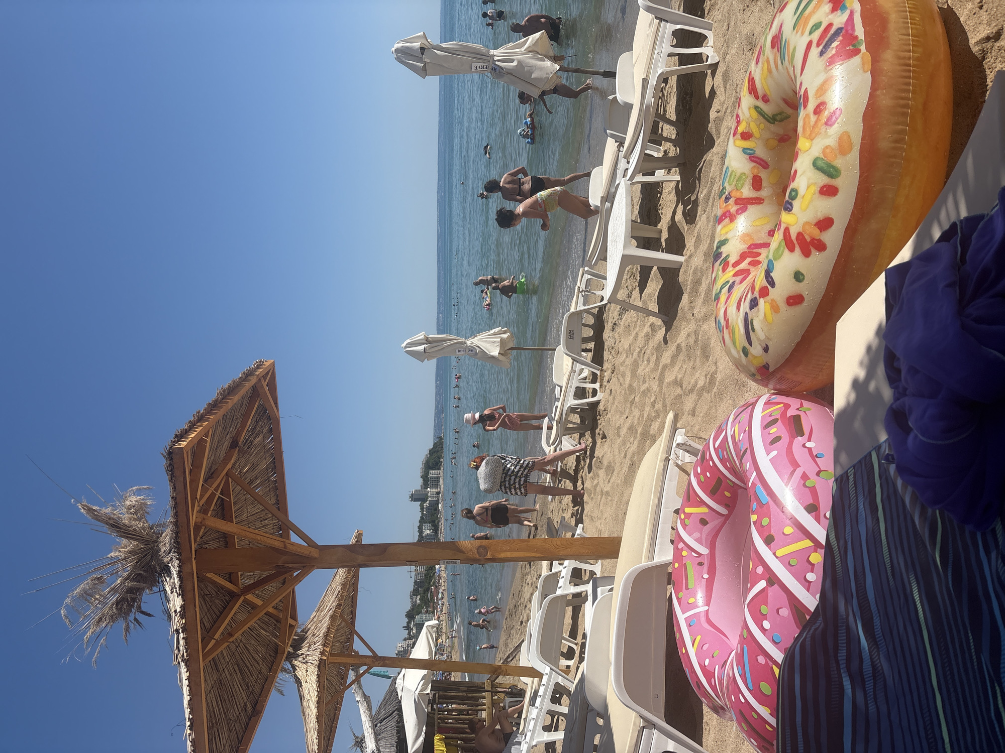 The image depicts a sunny day at a crowded beach, with the sea and distant city buildings in the background. In the foreground, two large inflatable donut-shaped pool floats are positioned on the sandy beach, one pink with white frosting and sprinkles, the other yellow with colorful sprinkles. Several rows of white lounge chairs and a few white beach umbrellas are set up on the sand. In the water, numerous people are swimming, wading, and some are using inflatable toys. The overall scene suggests a typical summer vacation atmosphere at a popular seaside resort.