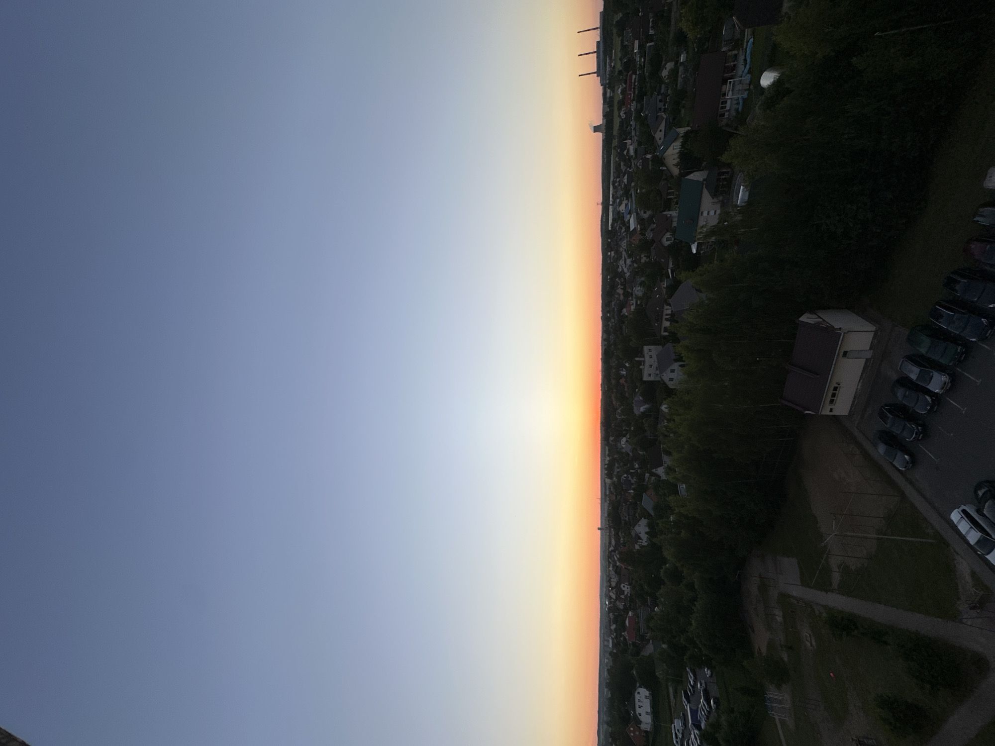This is a high-angle, wide-shot photograph of a suburban landscape in the Шчомысліцкі сельскі Савет, Belarus, during dusk. The sky is a gradient of clear blue at the top, transitioning to a soft orange and yellow horizon where the sun has set. Below the horizon, a dense area of trees and houses stretches across the middle ground. The foreground features a parking lot with several cars and a small, light-colored building with a dark roof. Further into the scene, a playground with metal structures is visible, alongside more trees and houses of varying sizes and roof colors. In the far distance, industrial structures, including what appear to be power plant cooling towers and smokestacks, are silhouetted against the twilight sky. The overall impression is a peaceful, residential area at the edge of an industrial zone as day transitions to night. There are no people visible in the frame.