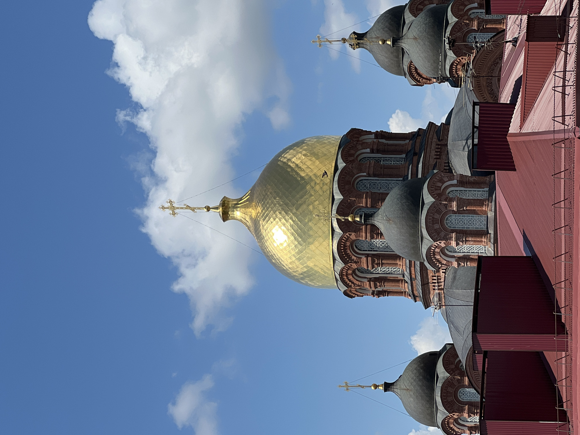This image shows a view of the domes of the Catherine Cathedral in Krasnodar, Russia. The cathedral features several prominent onion domes, one of which is gilded and reflective, while others are dark gray. The domes are adorned with crosses. The building appears to be constructed from red brick. In the foreground, there are red-colored rooftops and structures, suggesting an urban setting. The sky above is blue with scattered white clouds. There are no people visible in the image.