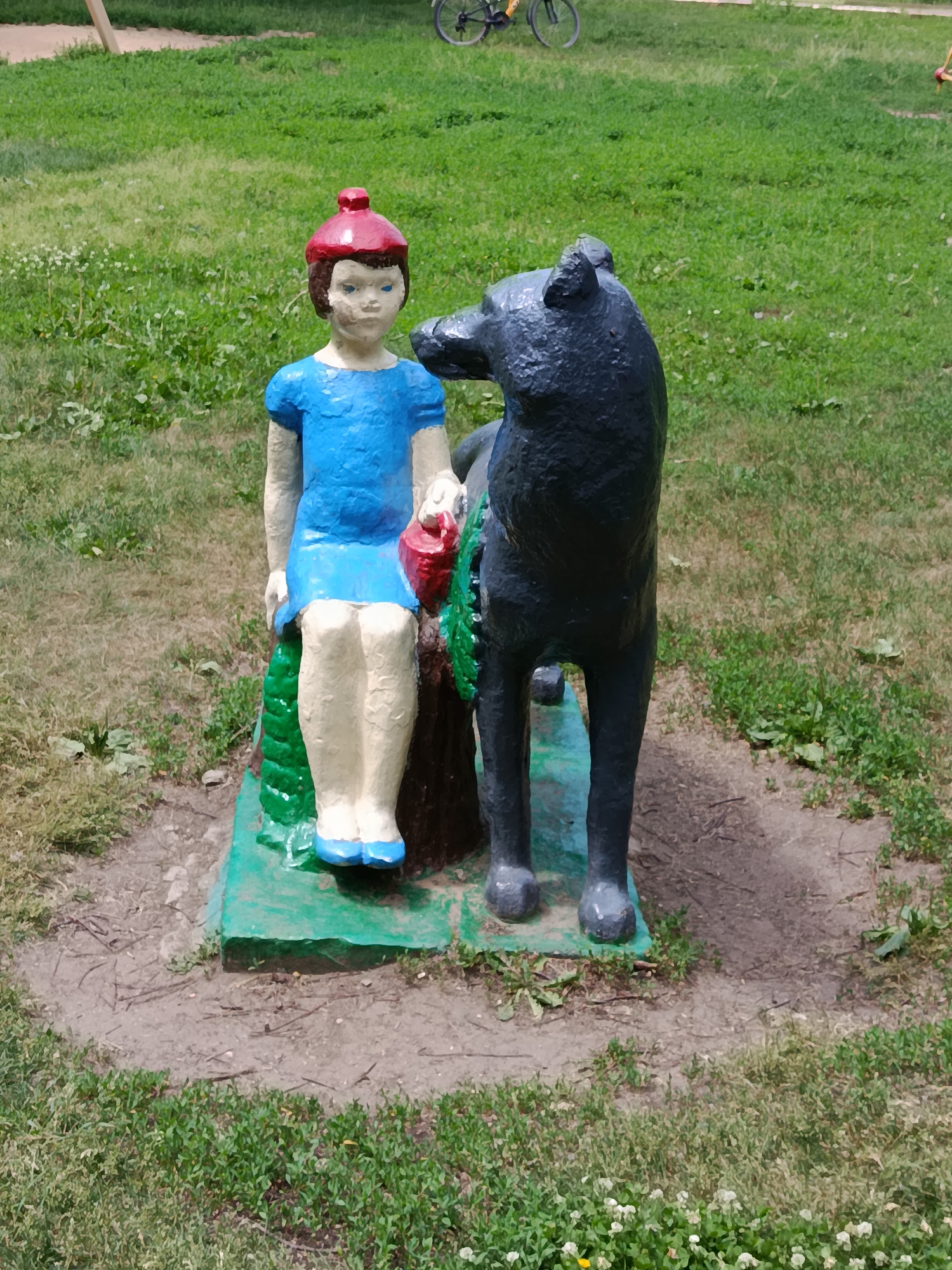 The image depicts a concrete sculpture of a young girl sitting beside a dog, positioned outdoors on a patch of grass. The girl is depicted wearing a blue dress and a red cap, with her legs extended. She is holding a red object in her right hand. The dog is a dark color and is standing next to the girl. They are situated on a green base. In the background, there is more green grass and a portion of a bicycle wheel is visible, suggesting the location is likely a park or recreational area. The scene appears to be in the city of Marx, Russia, based on the provided context.