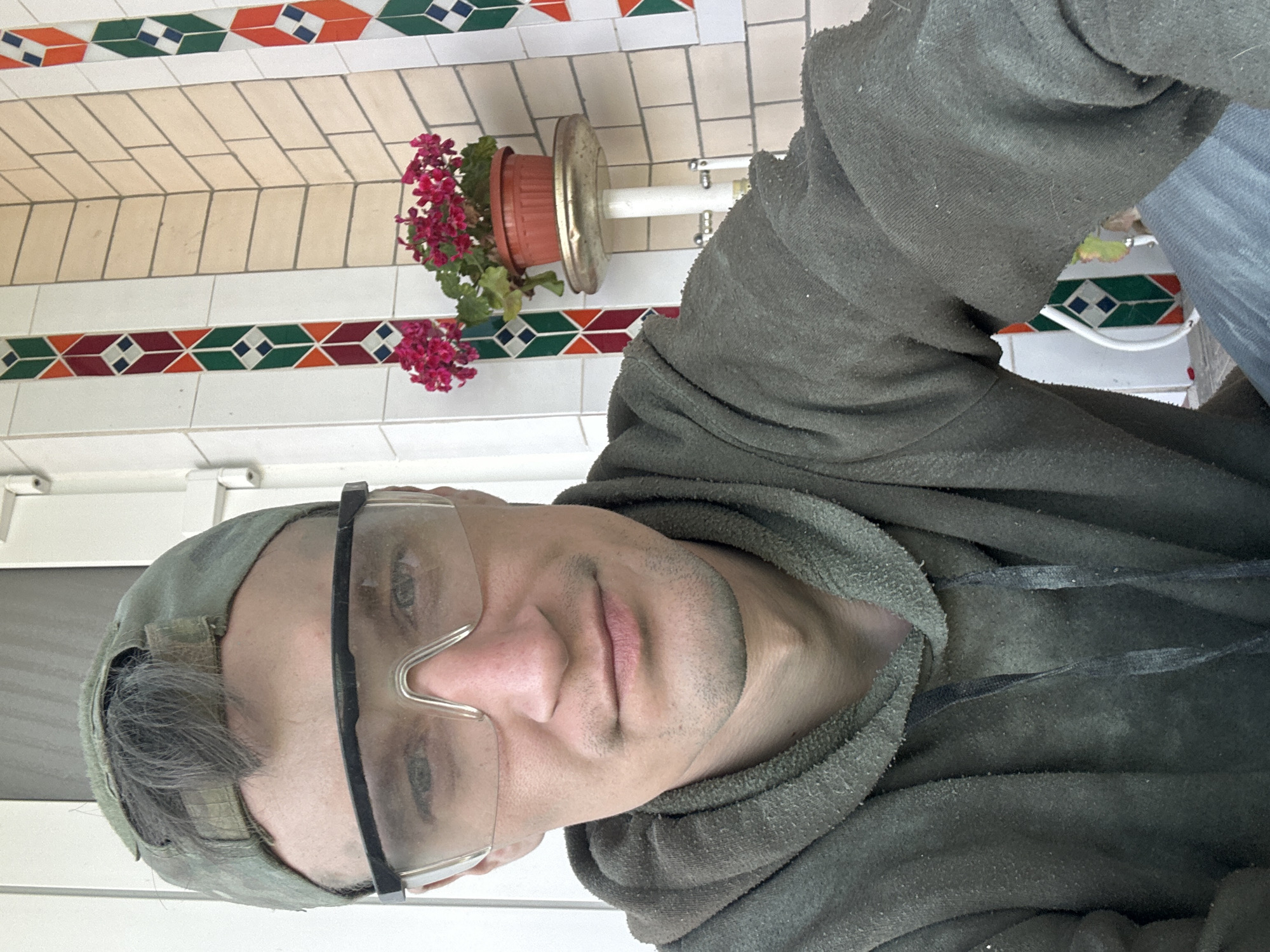 A man, wearing a camouflage baseball cap, clear safety glasses, and a dark green hooded sweatshirt, takes a selfie. He appears to be outdoors, possibly on a porch or patio, with a tiled wall and a potted plant visible in the background. The tiled wall features decorative geometric patterns in white, beige, green, orange, and dark blue. The man's face is the primary focus, with the rest of his body, including his torso and arms, also captured in the frame. The scene is likely located in Makiv, Ukraine, based on the provided location information.