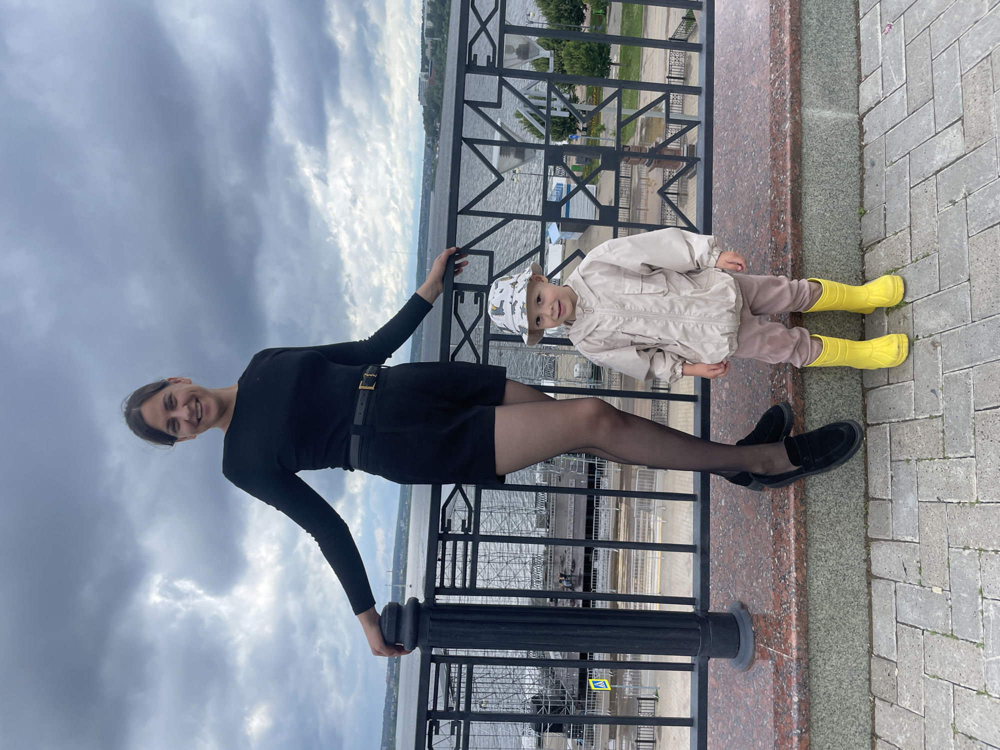 A woman and a young child stand side-by-side on a stone ledge, with a railing and a body of water behind them. The woman, dressed in a black long-sleeved shirt, black shorts, and black tights, leans on the railing with her left arm. She is smiling. The child, wearing a light-colored jacket, pink pants, and bright yellow boots, stands to her right, looking at the camera. The background shows a wide river, a bridge, and city buildings under a cloudy sky. The scene appears to be a waterfront promenade in Izhevsk, Russia.