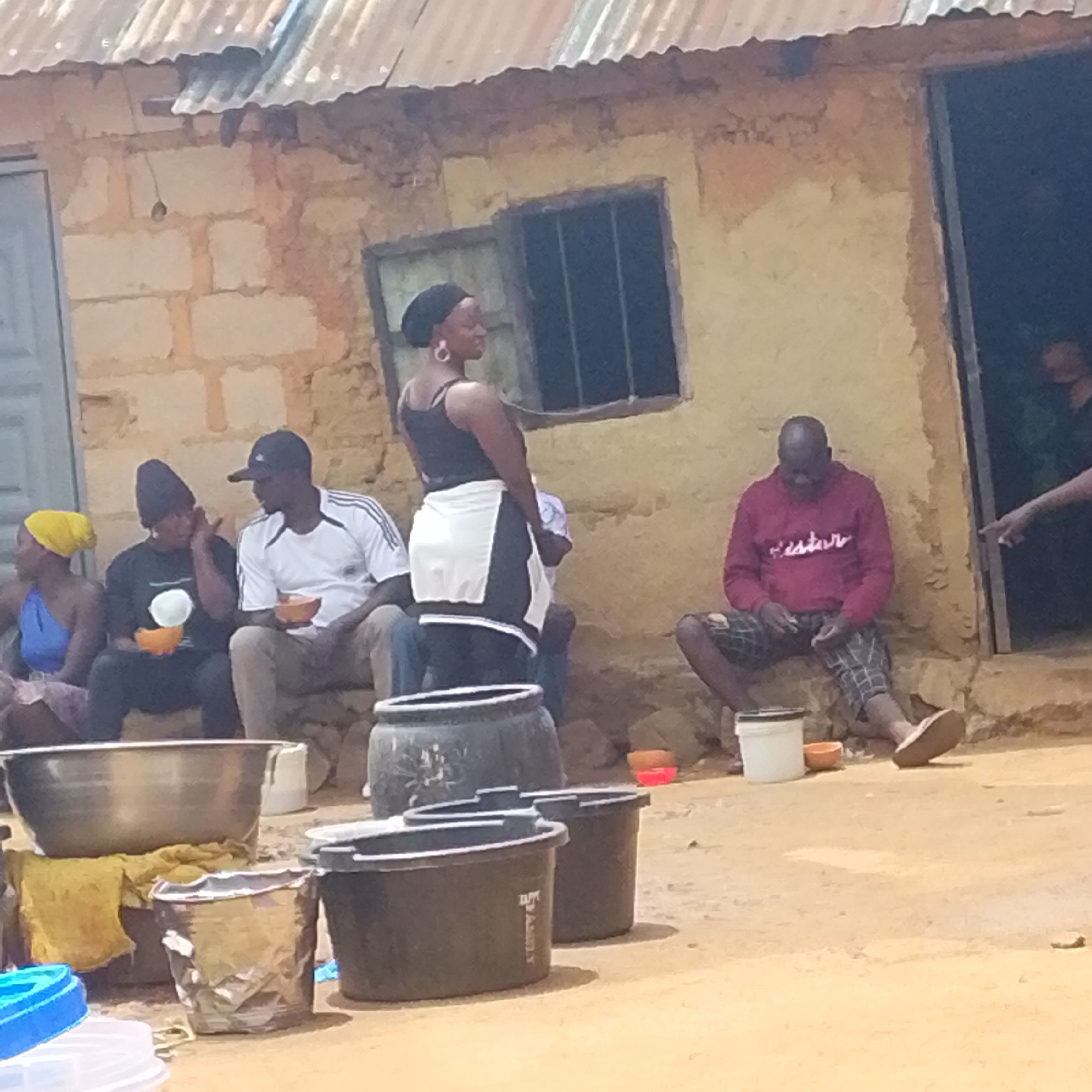 In an outdoor setting, possibly in a village or rural area, a group of individuals are gathered in front of a rustic building constructed of mud brick with a corrugated metal roof. The building has an open doorway and a window with bars. In the foreground, there are various containers, including large metal bowls, plastic tubs, and smaller buckets. Several people are seated on the ground or a raised step in front of the building. One woman in the center is standing and appears to be wearing a black tank top and a white apron-like garment. Another woman to her left wears a yellow headscarf and a blue top. A man to the woman's right, wearing a black beanie and a black t-shirt, is holding a small orange bowl. Further to the right, a man in a red hooded sweatshirt and plaid shorts is seated, looking down at his hands. A hand with a pointing finger is visible entering the frame from the right, near the doorway. The ground is dusty earth.