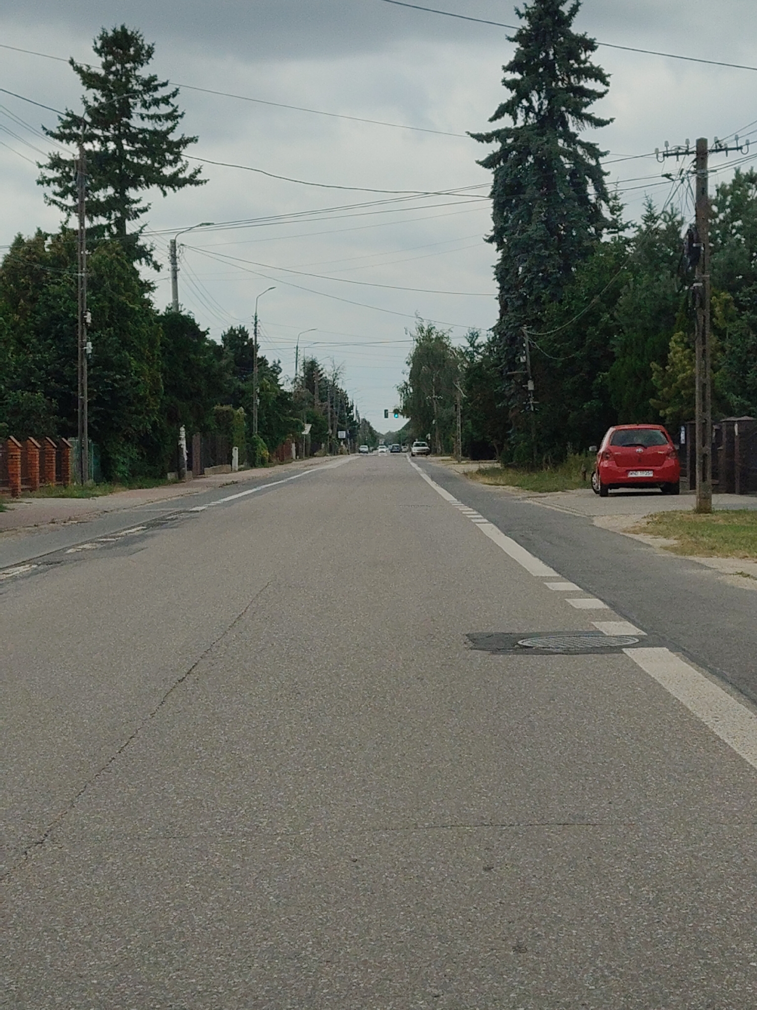 This is a medium-shot, eye-level, vertical photograph of a two-lane asphalt road in Łomna, Poland. The road runs from the foreground into the distance, with dense trees lining both sides. Power lines and streetlights are visible overhead. On the right side of the road, a red hatchback car is parked facing away from the camera. In the distance, a few other cars are visible on the road. The sky is overcast. The scene appears to be a suburban or rural area.