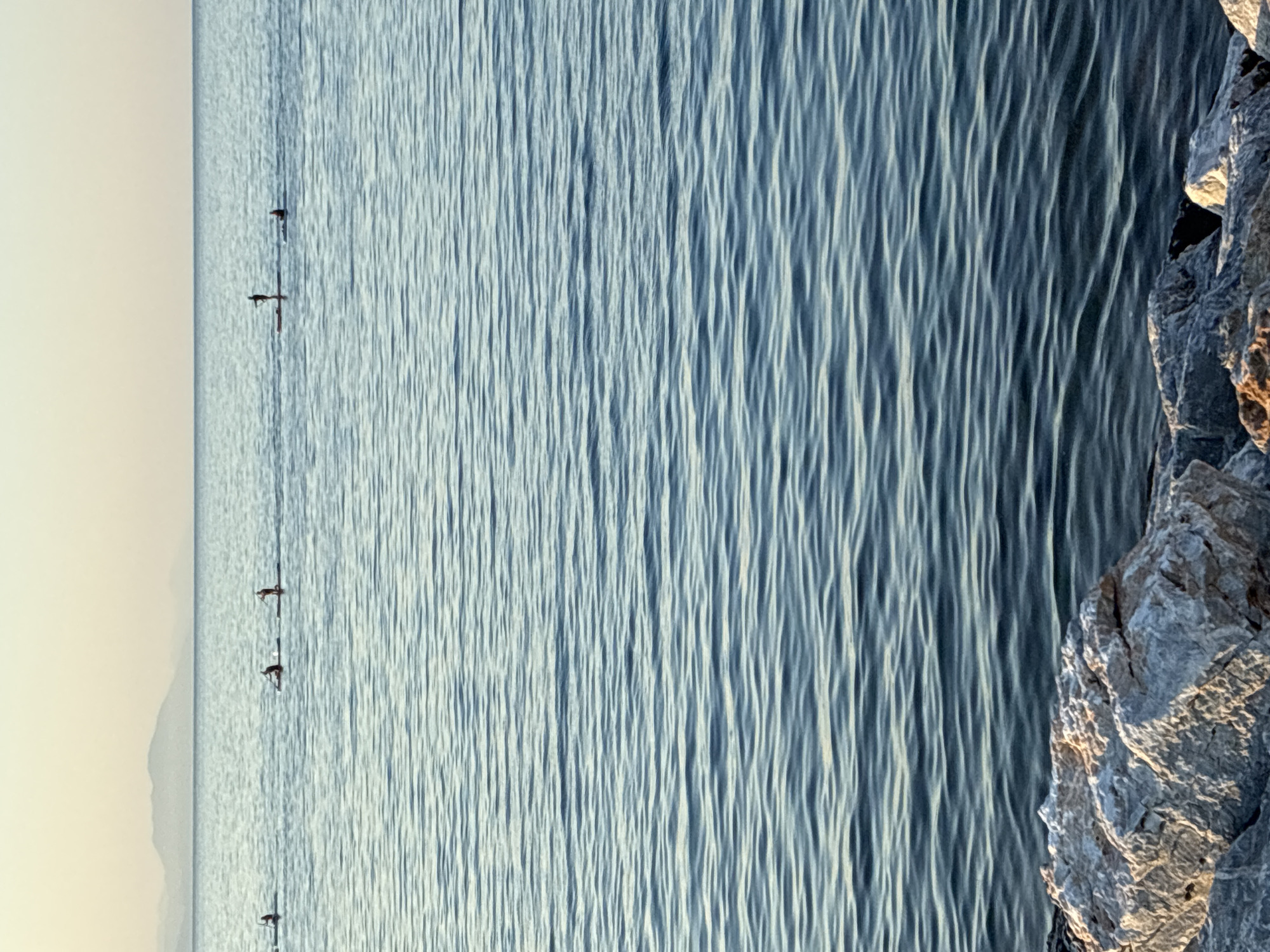 The image depicts a serene seascape with a line of paddleboarders in the distance, silhouetted against a pale sky and hazy mountains. The foreground features a rocky shoreline, with large, weathered stones in shades of gray and white. The sea itself is a calm expanse of blue, with gentle ripples reflecting the soft, diffused light of either sunrise or sunset. The paddleboarders are spaced out, each standing on their boards and holding paddles, engaged in a recreational activity on the water. Their small figures emphasize the vastness of the ocean and the tranquility of the scene. The overall impression is one of peaceful outdoor activity in a natural setting.