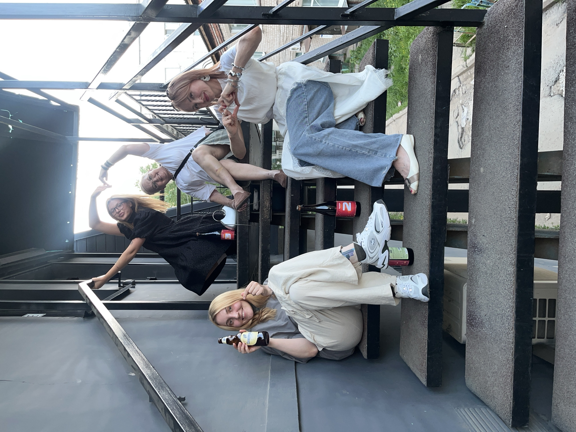 Captured in Chaikovsky, Russia, the photograph features four women posing on an outdoor staircase. The staircase has black metal railings and grey concrete steps. At the top, a woman with blonde hair, wearing a white shirt dress and blue jeans, is making a heart with her hands. To her lower left, another woman in a white shirt and olive-green shorts also makes a heart gesture. Adjacent to her is a woman in a black dress, also gesturing the heart shape. The fourth woman, in beige pants and a grey top, sits on the bottom step with a beer bottle. Multiple bottles of alcohol are on the stairs. The area around the staircase includes metal framework and glimpses of building exteriors.