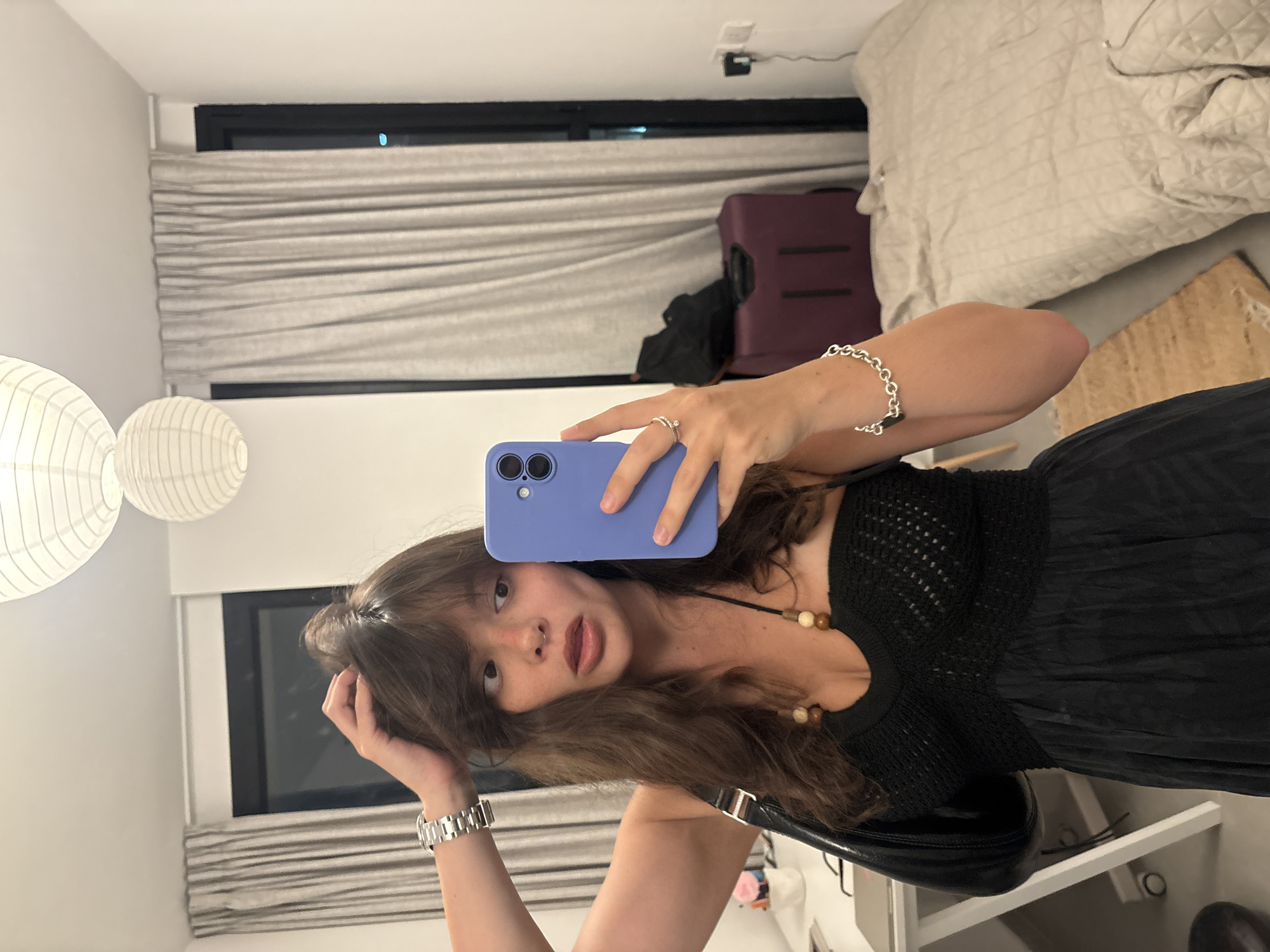 A young woman with long, wavy brown hair and bangs stands in a bedroom and takes a selfie in a mirror. She is wearing a black strapless dress with a crochet bodice and a silky skirt. She has a silver watch on her left wrist, a silver ring on her right index finger, and a silver bracelet with a heart-shaped charm on her right wrist. She is holding a purple smartphone in her right hand, which is angled to capture her reflection. Her left hand is raised and resting on her hair. Her mouth is slightly open, and she is looking to her left.
The room appears to be a modern hotel room or apartment. In the background, a large window is covered by beige curtains. To the right of the woman, a dark purple suitcase stands upright. A bed with a quilted cover is visible in the far right of the image. A desk with a white chair is on the left side of the room, with some items on it. Two white paper lanterns hang from the ceiling. The overall lighting suggests it is evening or indoors with artificial light. The presence of a modern suitcase and the interior design are consistent with a hotel room in Tel-Aviv, Israel.