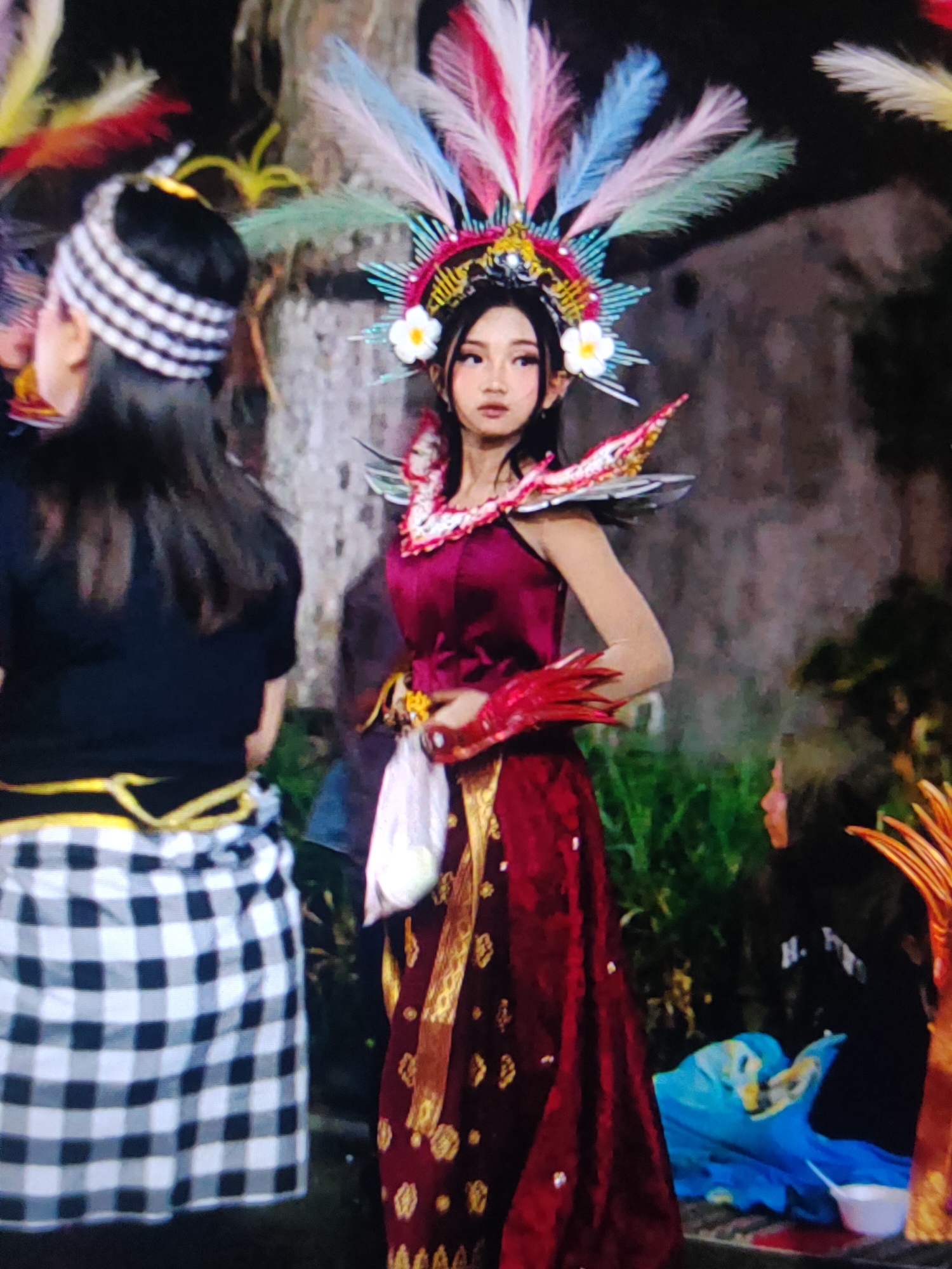 A young woman in a traditional maroon and gold dress and an elaborate headdress adorned with colorful feathers and flowers stands in the foreground, facing right. She is wearing red, claw-like gloves. To her left, another person, partially visible and facing away from the camera, wears a black shirt and a black and white checkered sarong tied with a yellow sash. The background is dark and appears to be an outdoor setting with some greenery. The woman's makeup includes prominent eyeliner and blush. The overall scene suggests a cultural performance or celebration. The location is Unknown, Unknown.