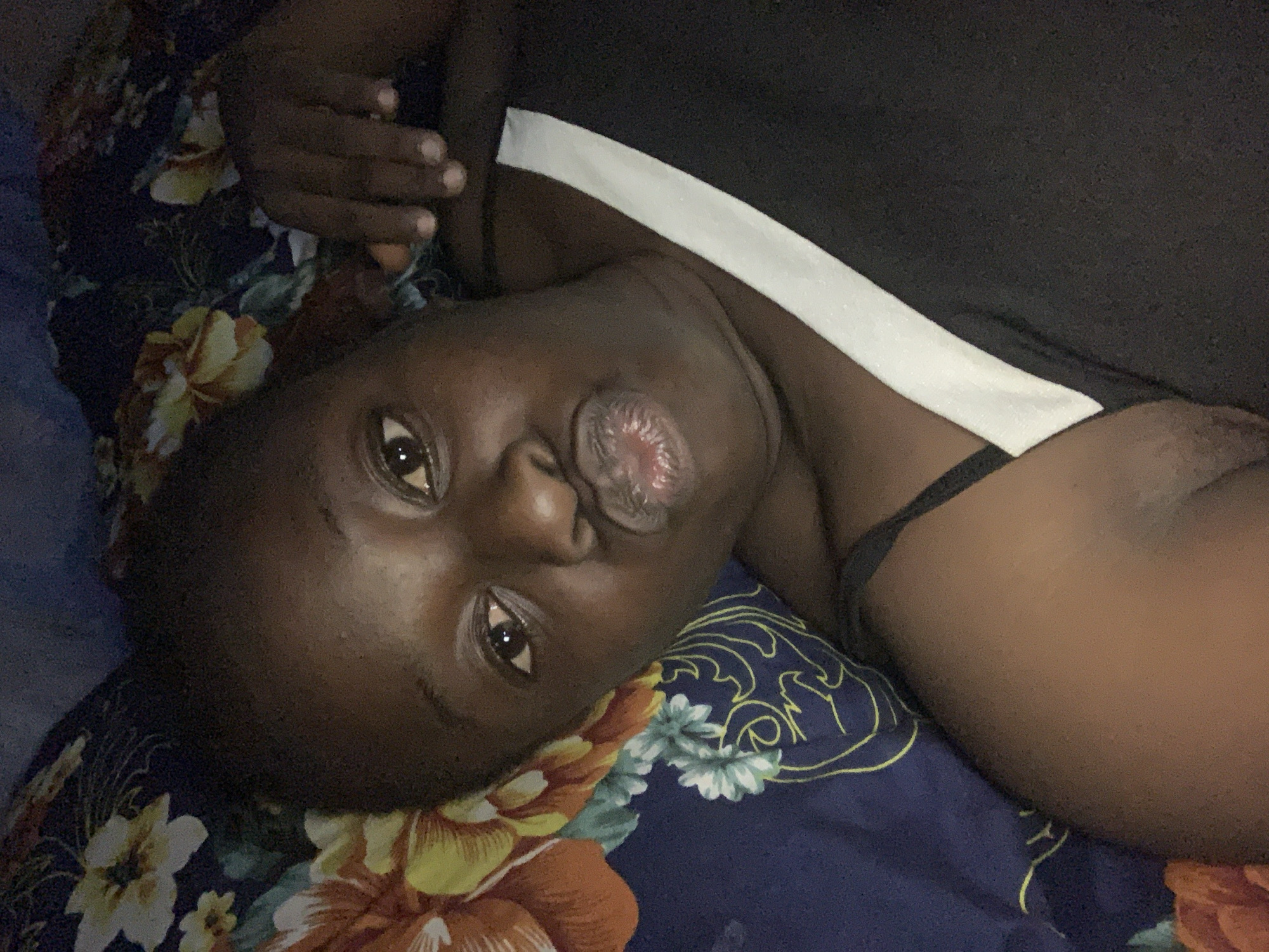 A person with dark skin is depicted in a close-up, upward-angled portrait, lying down and looking directly at the camera. Their head is tilted to the side, resting on a patterned bedspread. The individual's lips are pursed in a distinctive "duck face" or "fish-gaping" expression, revealing the inner surface of the lower lip. Their eyes are open, reflecting a light source, and convey a direct gaze.
The person is wearing a dark-colored top, possibly black, featuring a prominent white horizontal stripe across the upper chest or shoulder. A thin, dark strap, possibly from an undergarment or another layer of clothing, is also visible. The background is composed of a dark blue sheet or blanket adorned with large, vibrant floral patterns in orange, yellow, and light blue, interspersed with light blue abstract swirling lines.
The lighting in the image is dim, suggesting it was captured indoors during low-light conditions, likely at night or in the evening. The overall ambiance is relaxed and playful, indicating the person is posing for a photograph or selfie. There is no visible text within the image. The location context for this scene is Kukum, Nigeria.