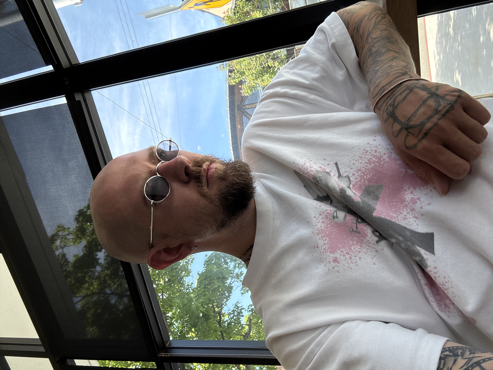 A low-angle, close-up photograph depicts a bald man with a beard and sunglasses, seated indoors beneath a glass roof. His left arm, covered in tattoos, is visible, resting on a white t-shirt with a graphic design that includes pink splatter. The man is looking toward the camera with a neutral expression. The roof structure is made of dark metal and glass panels, partially covered by a retractable sunshade. Through the glass, a view of trees and a blue sky is visible, along with a yellow street sign. The photo was taken in Los Angeles, United States of America.