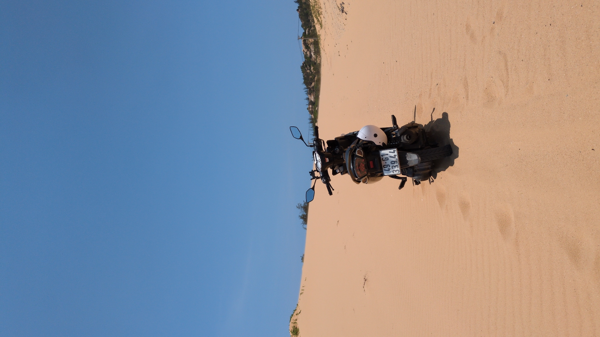 A motorcycle with license plate "49-G1 339.44" rests on sand dunes under a clear blue sky, potentially in the vicinity of Phuoc Dinh, Vietnam. A white helmet sits atop the motorcycle. The landscape includes vegetation and possibly distant structures on the horizon.
