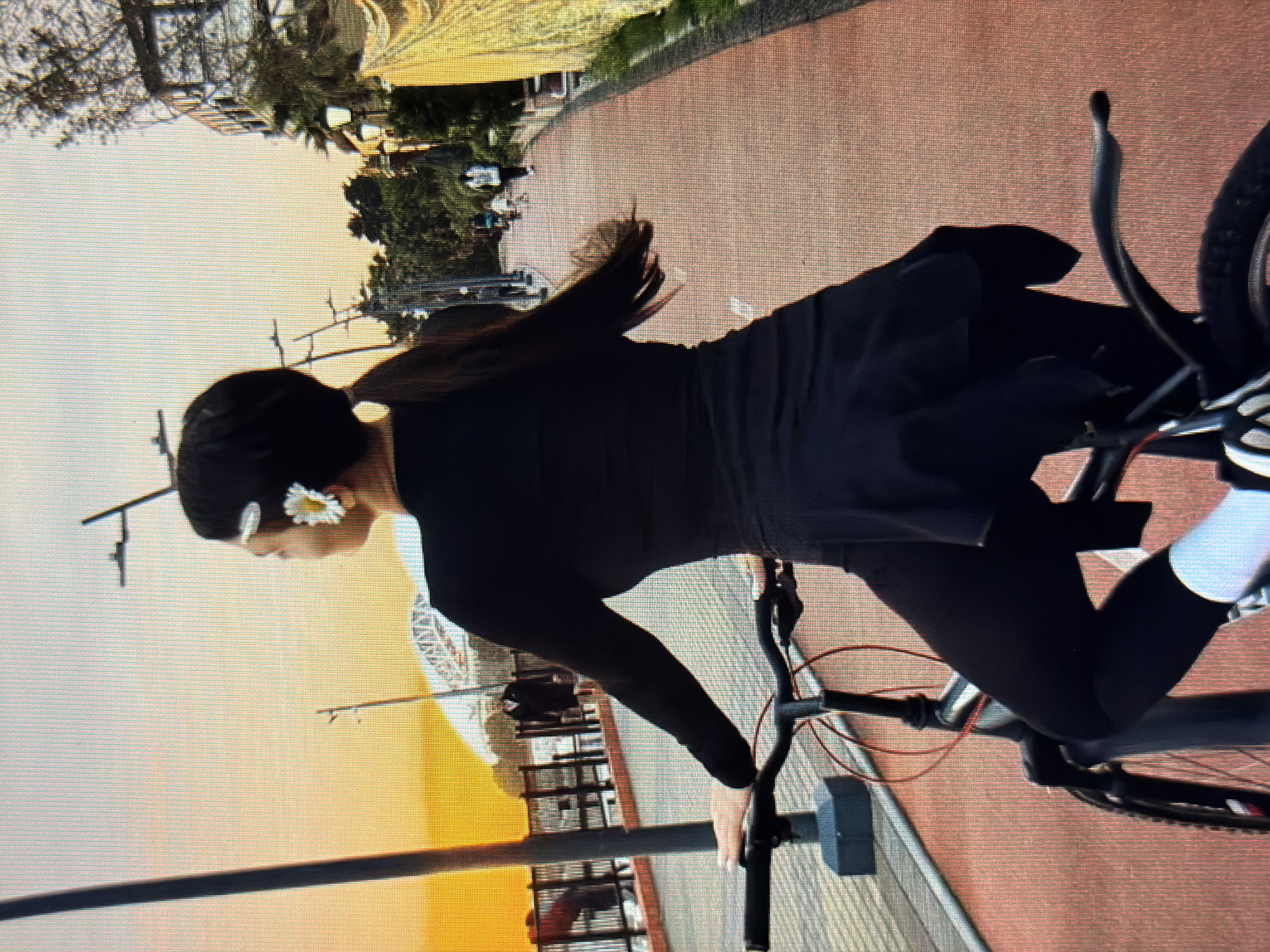 A woman with dark hair, adorned with a white flower clip, is captured from behind, seated on a bicycle. She is wearing a black long-sleeved top and black leggings, with white socks visible. The bicycle is dark-colored with red accents on its frame. The woman is holding the handlebars, appearing to be in motion. The scene is set on a paved pathway, possibly a promenade, with a red surface and a railing to the left. In the background, a bridge structure is visible, and beyond that, a bright orange sky suggests either sunrise or sunset. The environment appears to be urban, with lampposts and trees lining the pathway. The city of Sirius, Russia, is not depicted in this image.