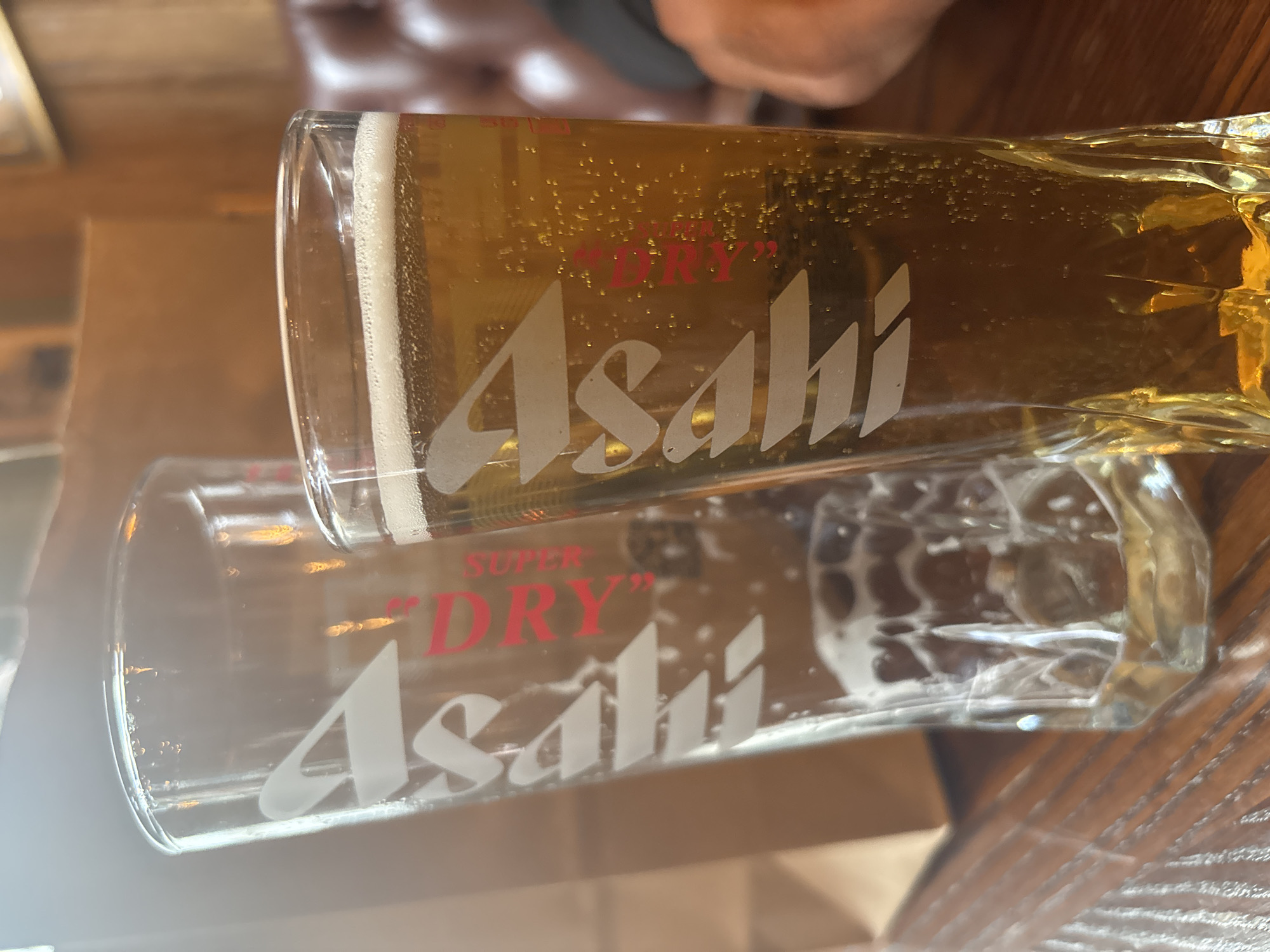 The image shows two Asahi beer glasses, one filled with beer and one empty, likely resting on a wooden table. The beer in the filled glass is light in color with visible bubbles and a frothy head. The glasses feature the "SUPER 'DRY'" Asahi logo in red and white. The background is blurred and appears to be the interior of a pub or restaurant with wooden paneling and some indistinct figures. There is no information to place this image in London, UK.