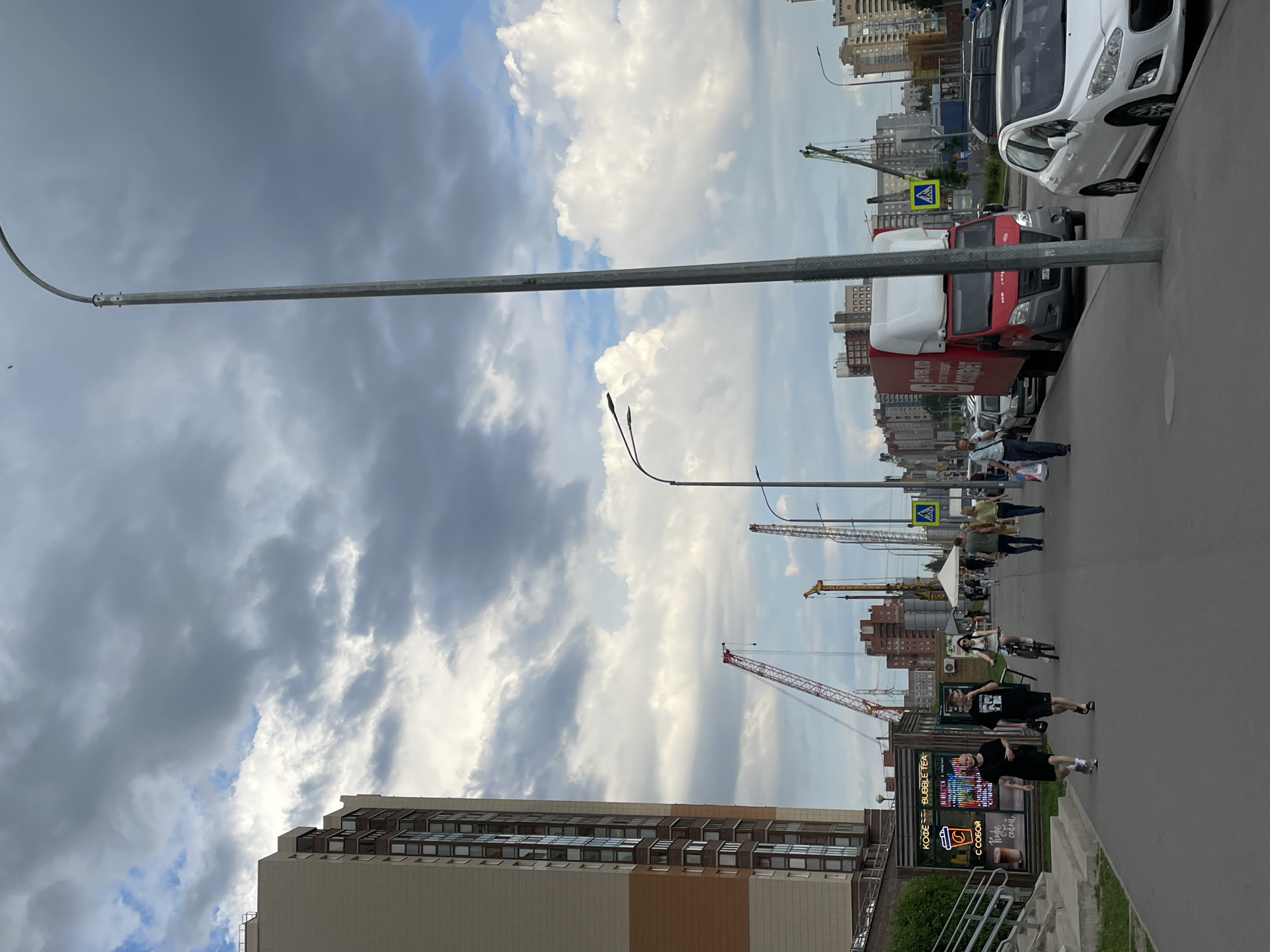 The image presents a daytime urban street scene, likely in Saint Petersburg, Russia, characterized by a mix of pedestrians, vehicles, and construction activity under a cloudy sky. The perspective looks down a street lined with buildings.
The sky is largely overcast with significant grey cloud cover, although patches of brighter blue sky and white clouds are visible, suggesting variable weather.
On the left side of the street, a prominent modern building with a multi-story, beige and brown facade featuring numerous windows stands tall. In front of this building, closer to the viewer, is a large, illuminated billboard. The billboard displays Cyrillic text: "КОФЕ BUBBLE TEA" (Coffee Bubble Tea) and "С СОБОЙ" (To Go), along with a graphic of a coffee cup. Several people are walking on the sidewalk in this area, including two individuals in dark clothing near the billboard. Further down the street, a person is seen riding a bicycle.
A long, gray street lamp arm extends horizontally across the upper portion of the frame, with another lamp visible in the distance.
On the right side of the street, two vehicles are parked: a white hatchback car (resembling a Peugeot) and behind it, a red and white truck. Some Cyrillic text is faintly visible on the side of the truck. A yellow, triangular pedestrian crossing sign is positioned behind the parked vehicles. Multiple pedestrians are walking on the sidewalk on this side, with some nearing the crosswalk.
In the background, numerous urban buildings are visible, and several large construction cranes – one yellow and one red – dominate the skyline, signifying active development in the area. The overall scene depicts a busy, evolving city environment during what appears to be late morning or afternoon.