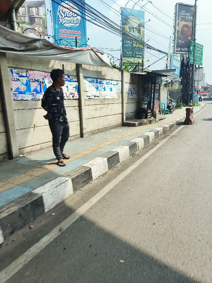 The image shows a man standing on a sidewalk in front of a concrete wall. There is a bus stop shelter behind him and a small shop with a red sign on it. He is wearing a dark shirt and black pants and black shoes. A row of large advertisements are on the wall behind him. The wall is covered in graffiti. The sidewalk is paved with black and white tiles. There is a street in front of the sidewalk. The street is paved with asphalt. There are several power lines running across the sky. The street is empty of cars. The man is looking at the street. The location is at the coordinates -6.852, 107.498 in Padalarang, Indonesia.