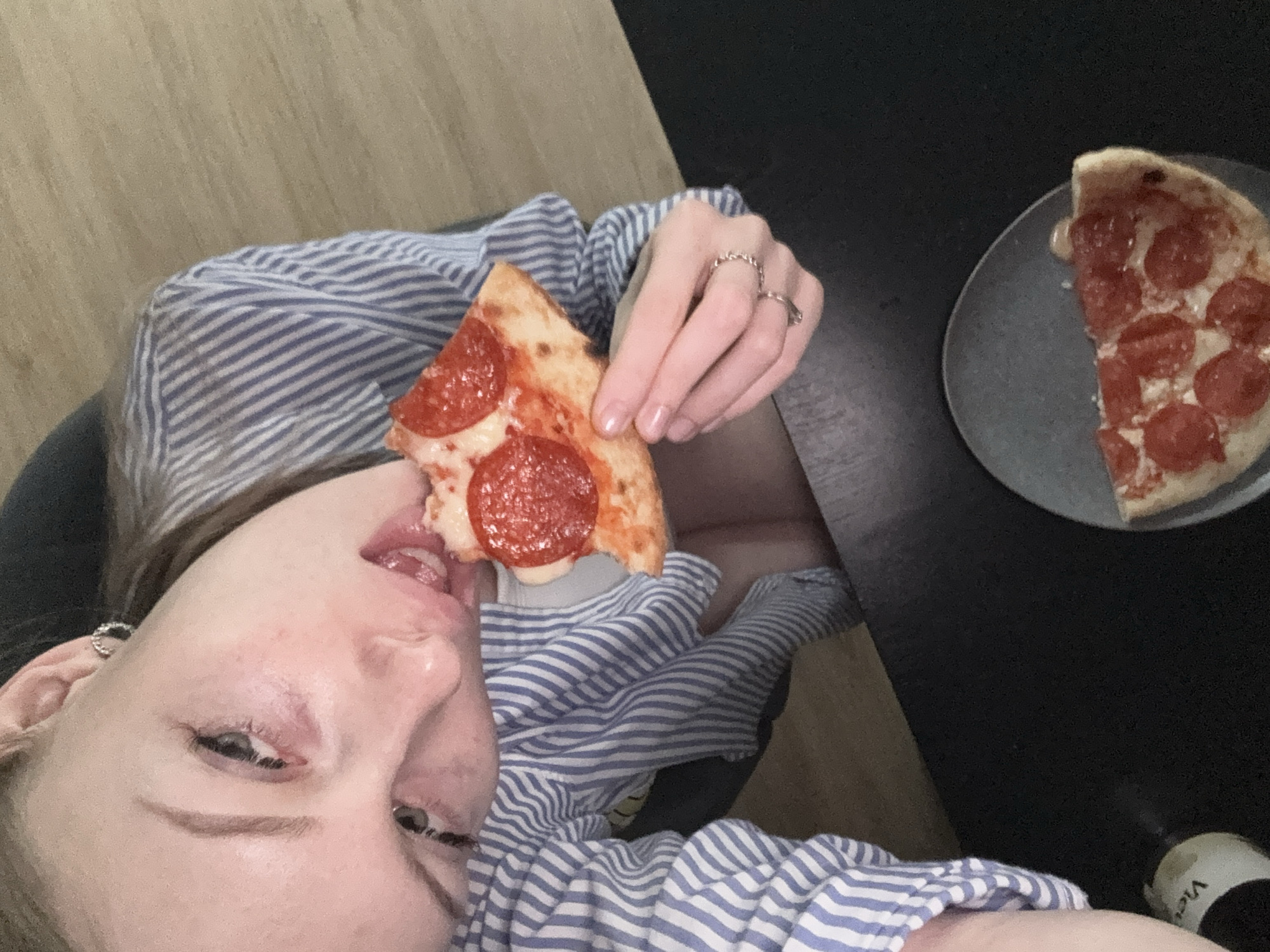 A woman with fair skin, wearing a blue and white striped shirt, is eating a slice of pepperoni pizza. She is holding the slice with her right hand. A half-eaten pizza sits on a gray plate to the right of the woman on a black table. A bottle with white and black writing is visible in the bottom right corner. The background includes a tan wall and a wooden surface. The location is not discernible as Perm, Russia from the visual information provided.