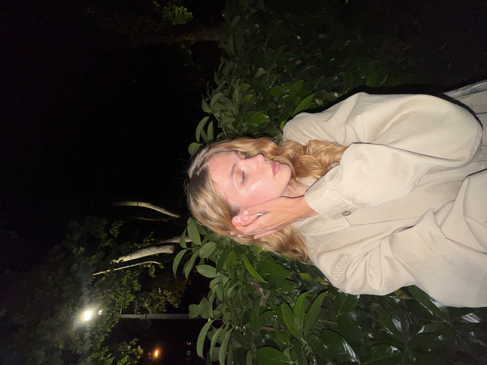 A woman with blonde hair, wearing a light beige jacket, rests her head in her hands. She is near a leafy bush or small tree. The background is dark, suggesting nighttime. In the distance, there's a street light and another smaller light source. The location is identified as Tbilisi, Georgia.