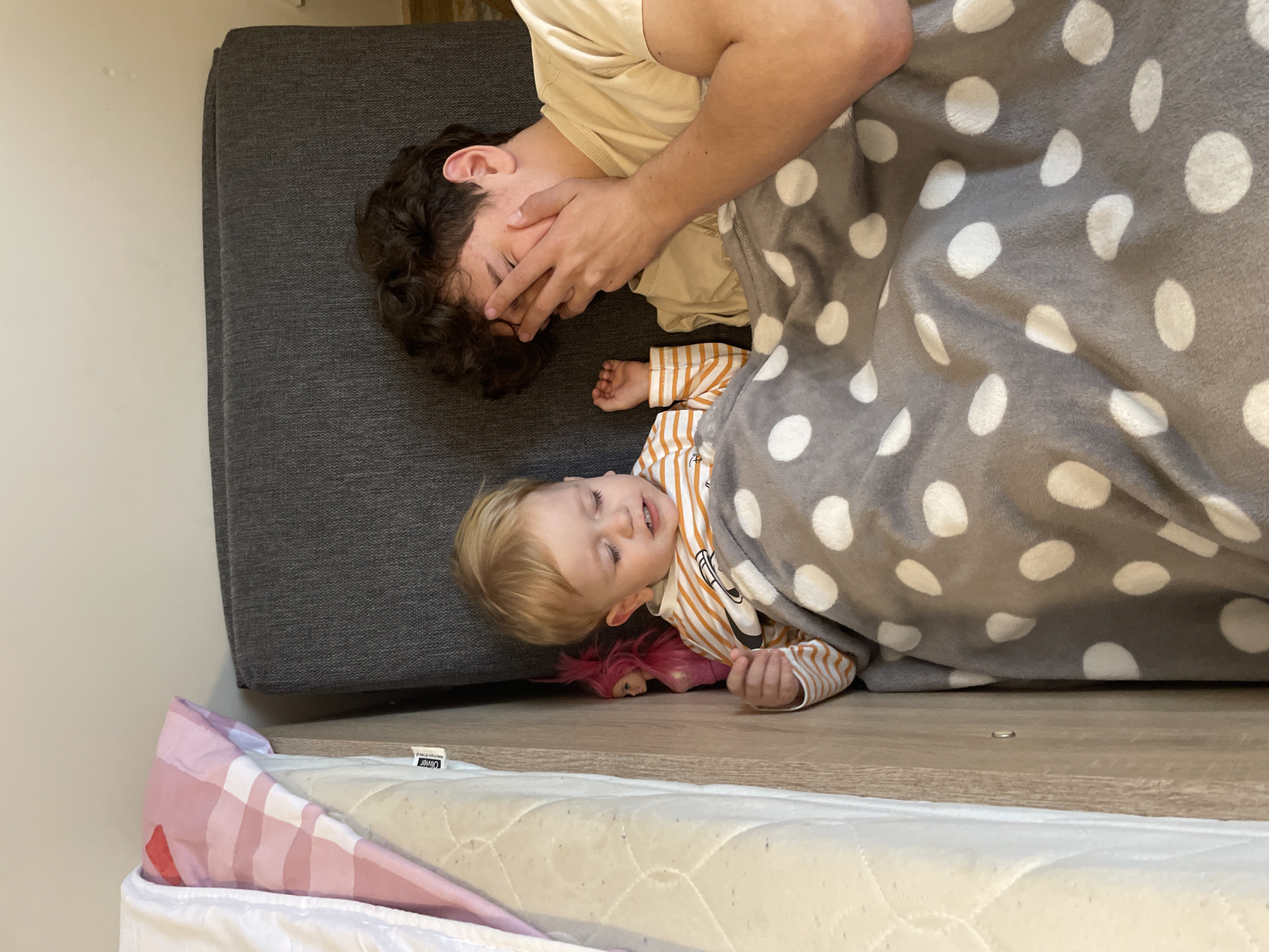 The image captures an indoor scene in Unknown, Unknown, featuring two individuals, a young child and an older person, lying under a grey blanket patterned with white polka dots. The child, positioned on the left, has blonde hair, wears an orange and white striped long-sleeved shirt, and holds a pink-haired doll in their right hand. Their eyes are partially closed. To the right, the older individual, characterized by dark, curly hair and a light-colored t-shirt, lies with their right hand covering their face. To the far left, a cream-colored mattress with a visible "Olivier" tag is partially seen, topped with a folded pink and white checkered comforter. A narrow light wood panel separates the mattress from the child. A dark grey upholstered surface is visible behind the older individual, and a light-colored wall forms the upper background.
