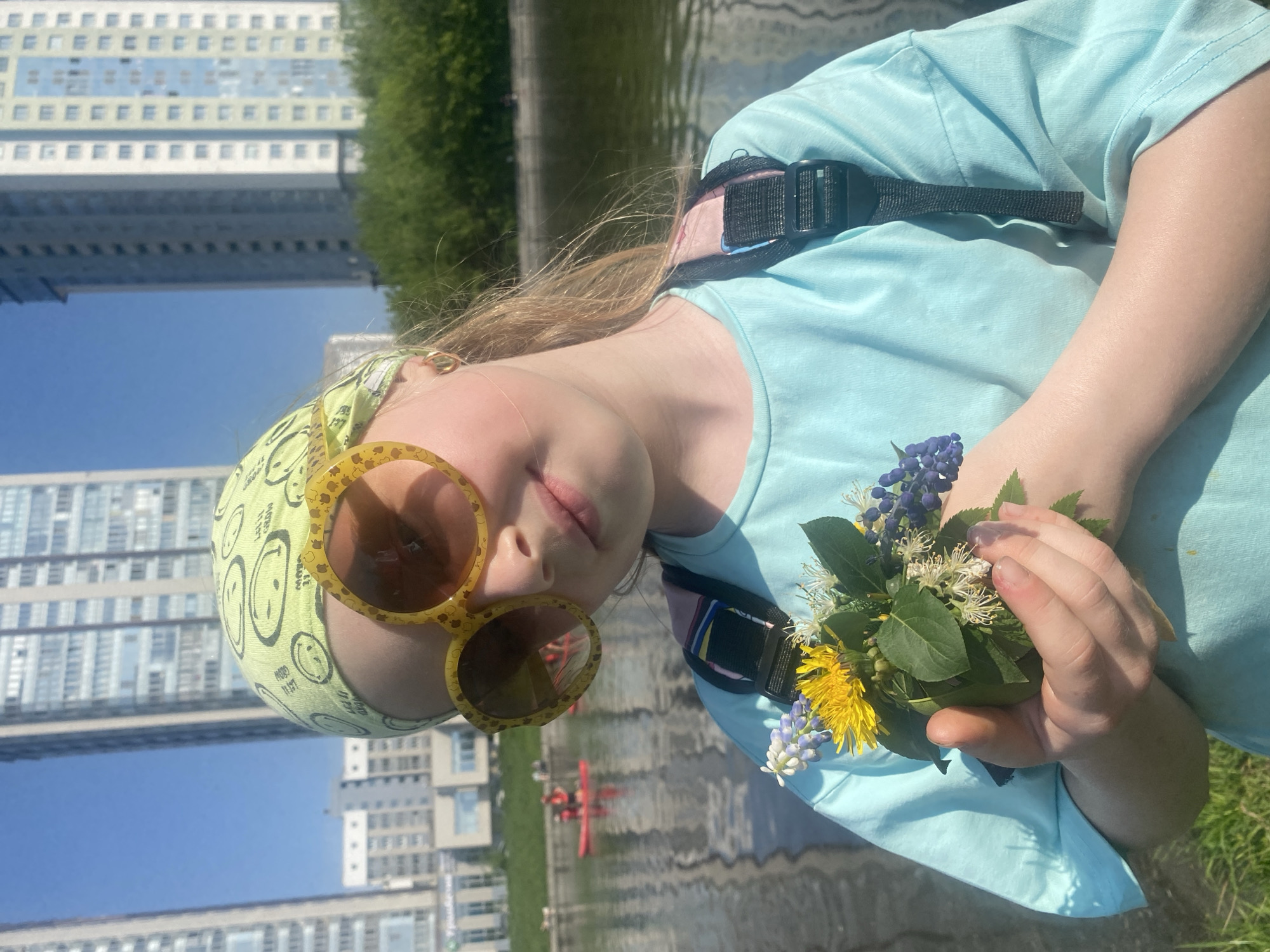 A young girl with blonde hair is holding a small bouquet of wildflowers in her hands. She is wearing sunglasses and a headscarf. She is wearing a turquoise t-shirt with backpack straps. The background shows a body of water and several modern buildings. The body of water is the Iset River and the buildings are part of the Yekaterinburg City complex in Yekaterinburg, Russia.